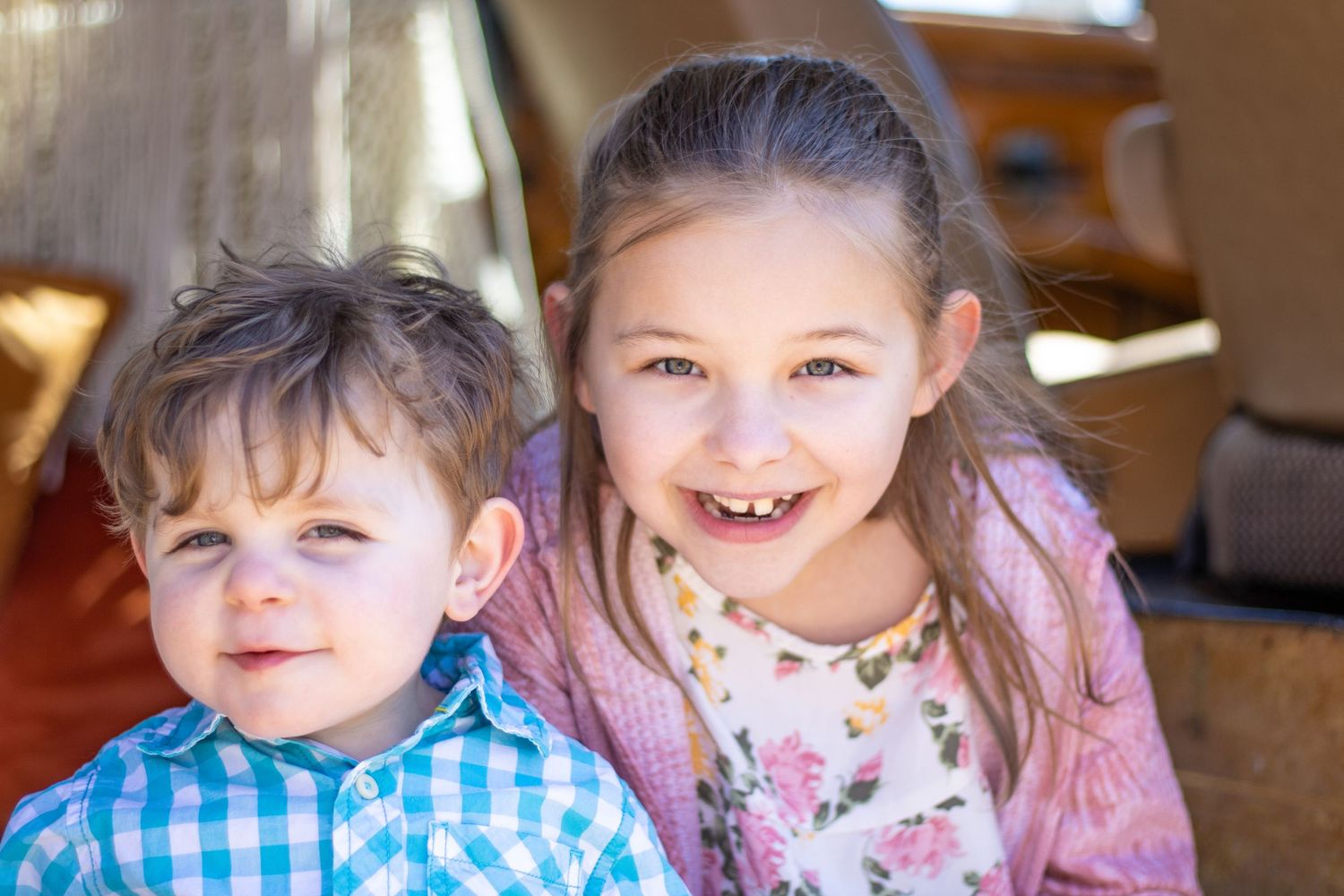 Two young children smiling together indoors, one wearing plaid and one in pink.