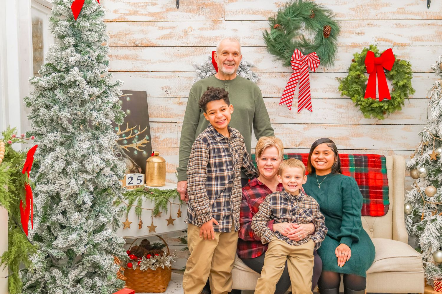 A family group poses beside a flocked Christmas tree and holiday decorations on a white wooden backdrop.