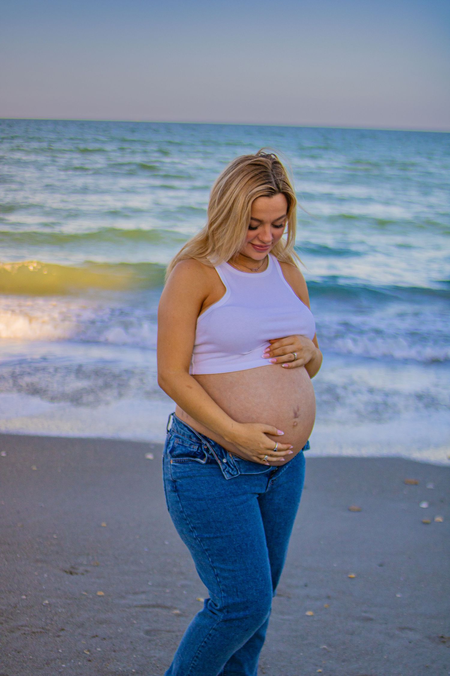 Pregnant woman in white top and jeans poses on beach at sunset.