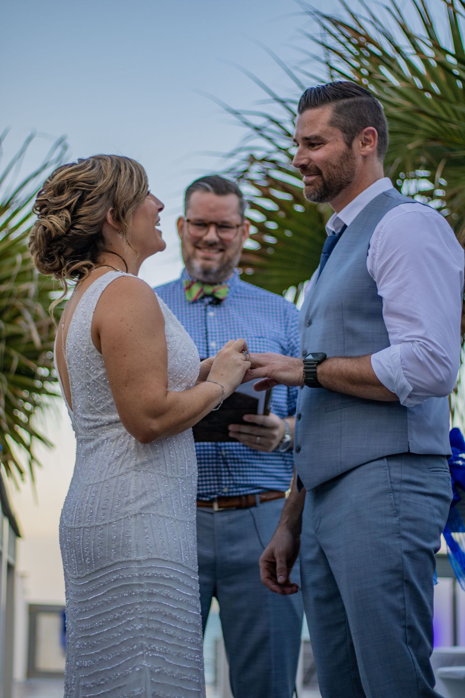 Couple exchanges rings during beachside wedding ceremony with officiant in background.