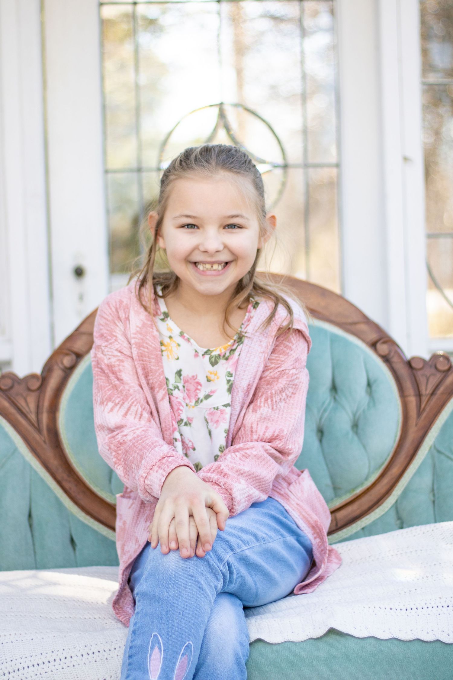 A child wearing pink cardigan and jeans sits on tufted turquoise chair with decorative window behind.