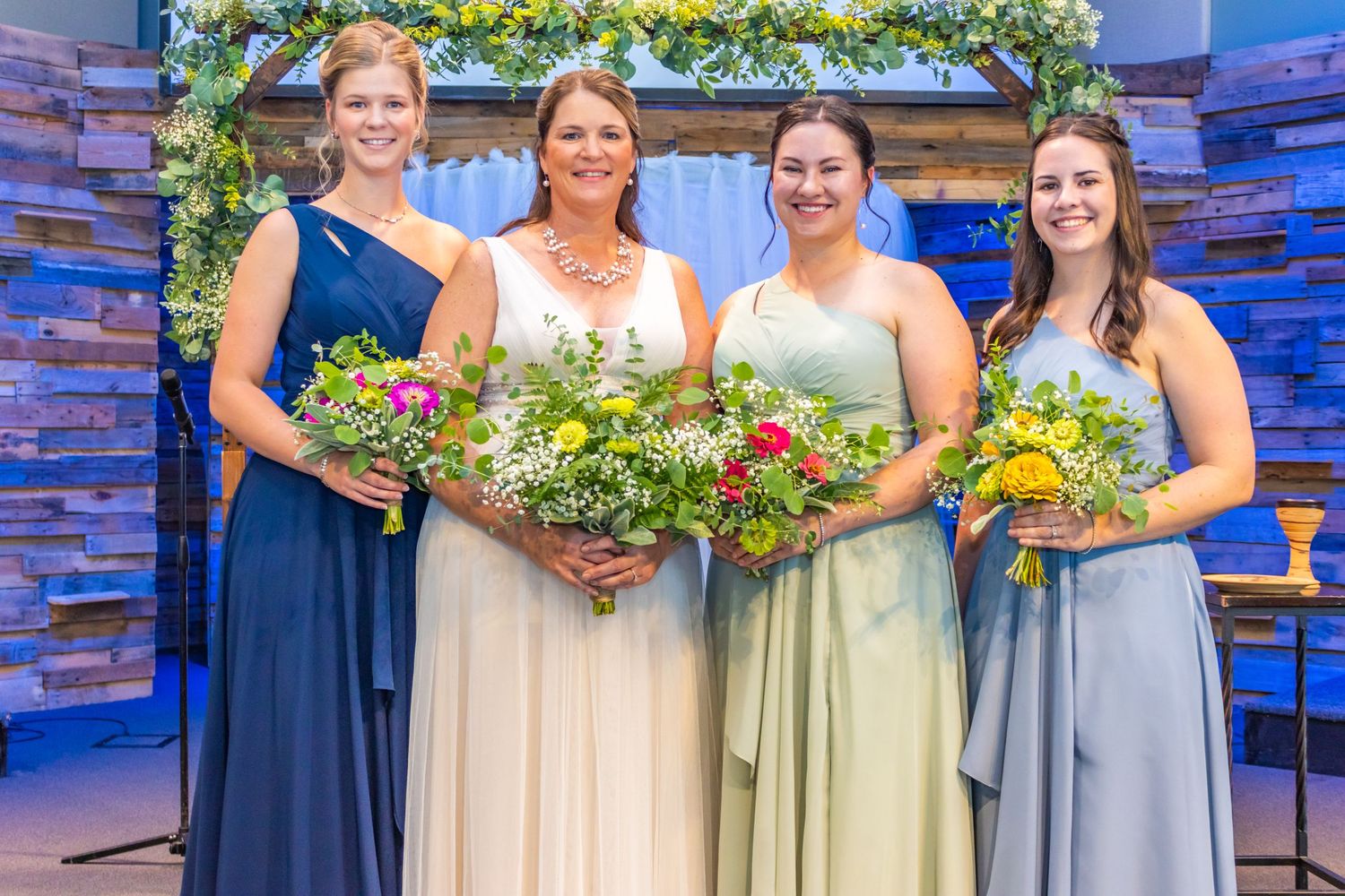 Wedding party poses together in coordinating blue and light colored dresses holding vibrant floral bouquets.