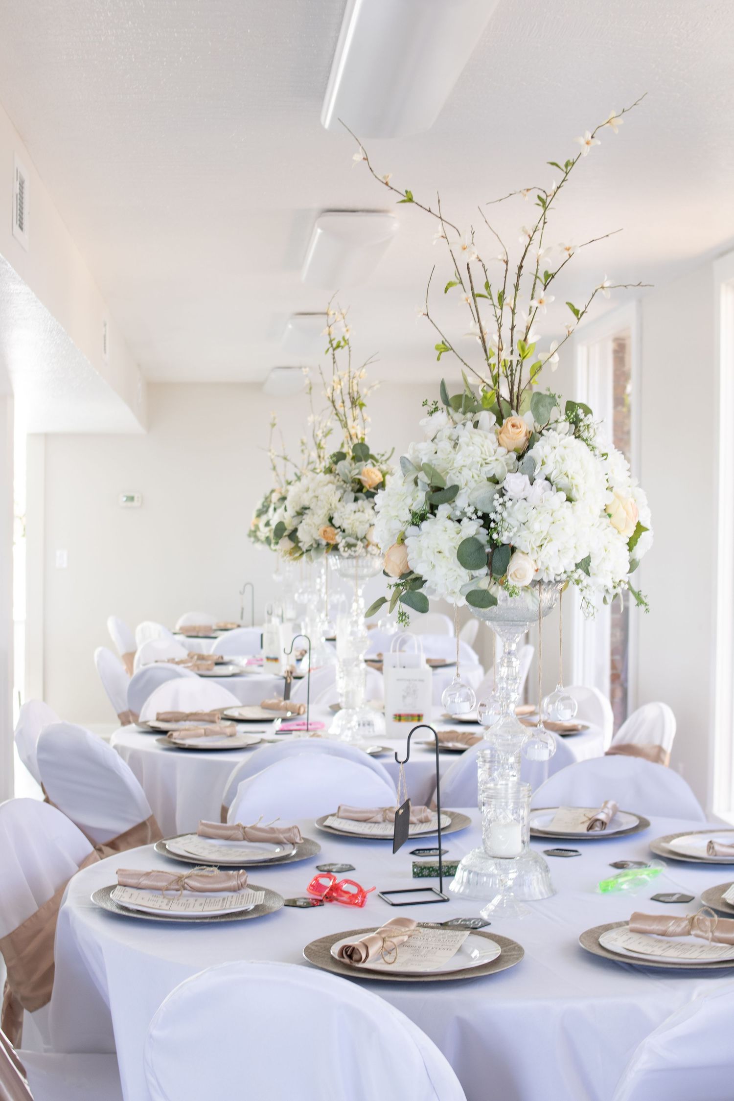 Elegant white wedding reception table setting with tall floral centerpiece and covered chairs.