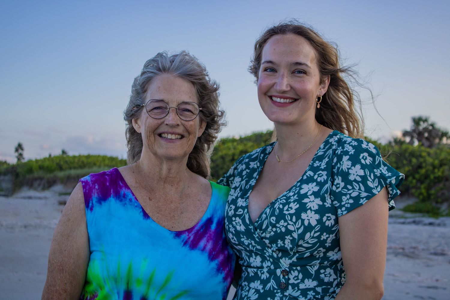 Two people share a warm moment at dusk on a beach with natural vegetation in the background.