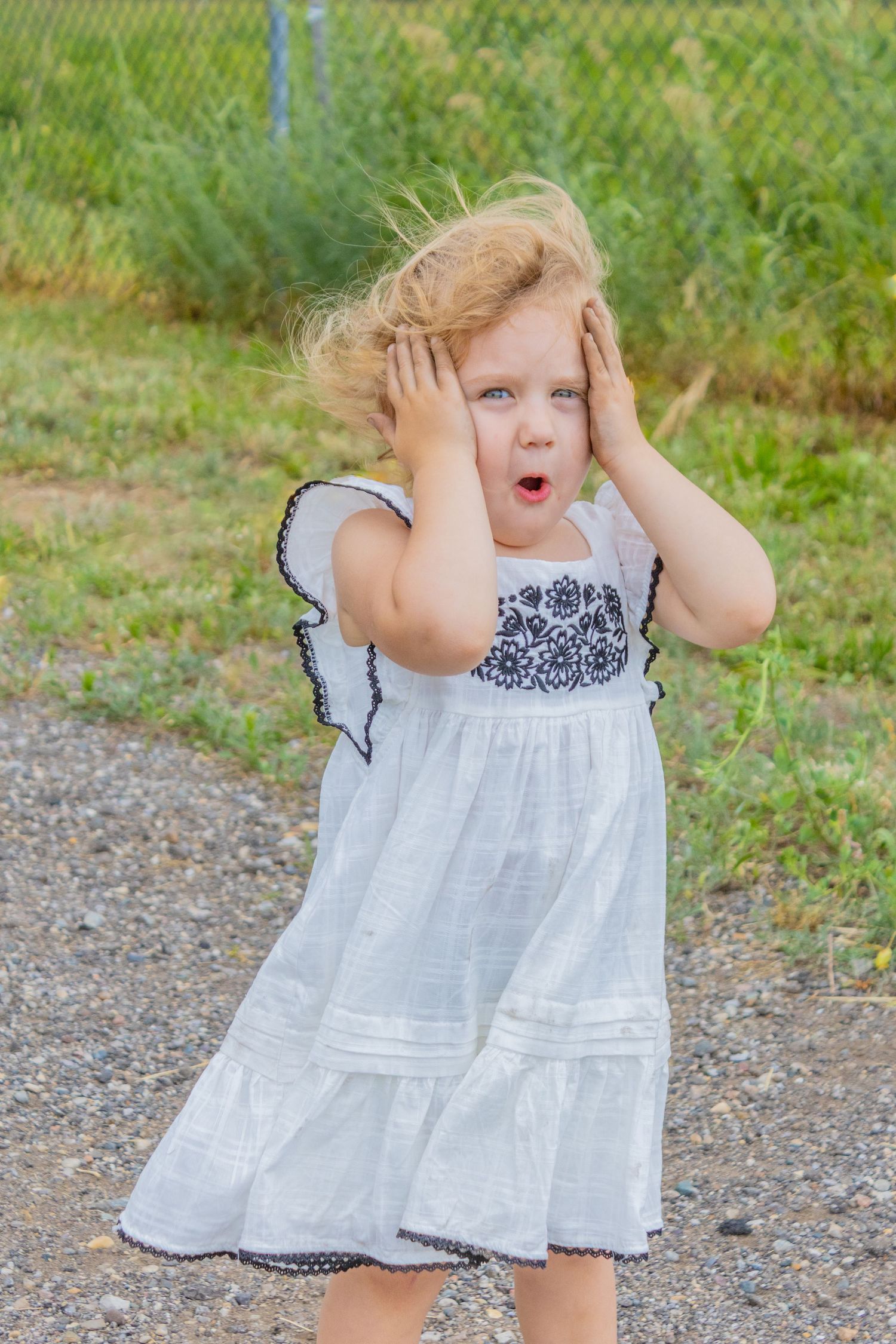 A young child in a white dress with black trim reacts expressively while covering their ears outdoors.