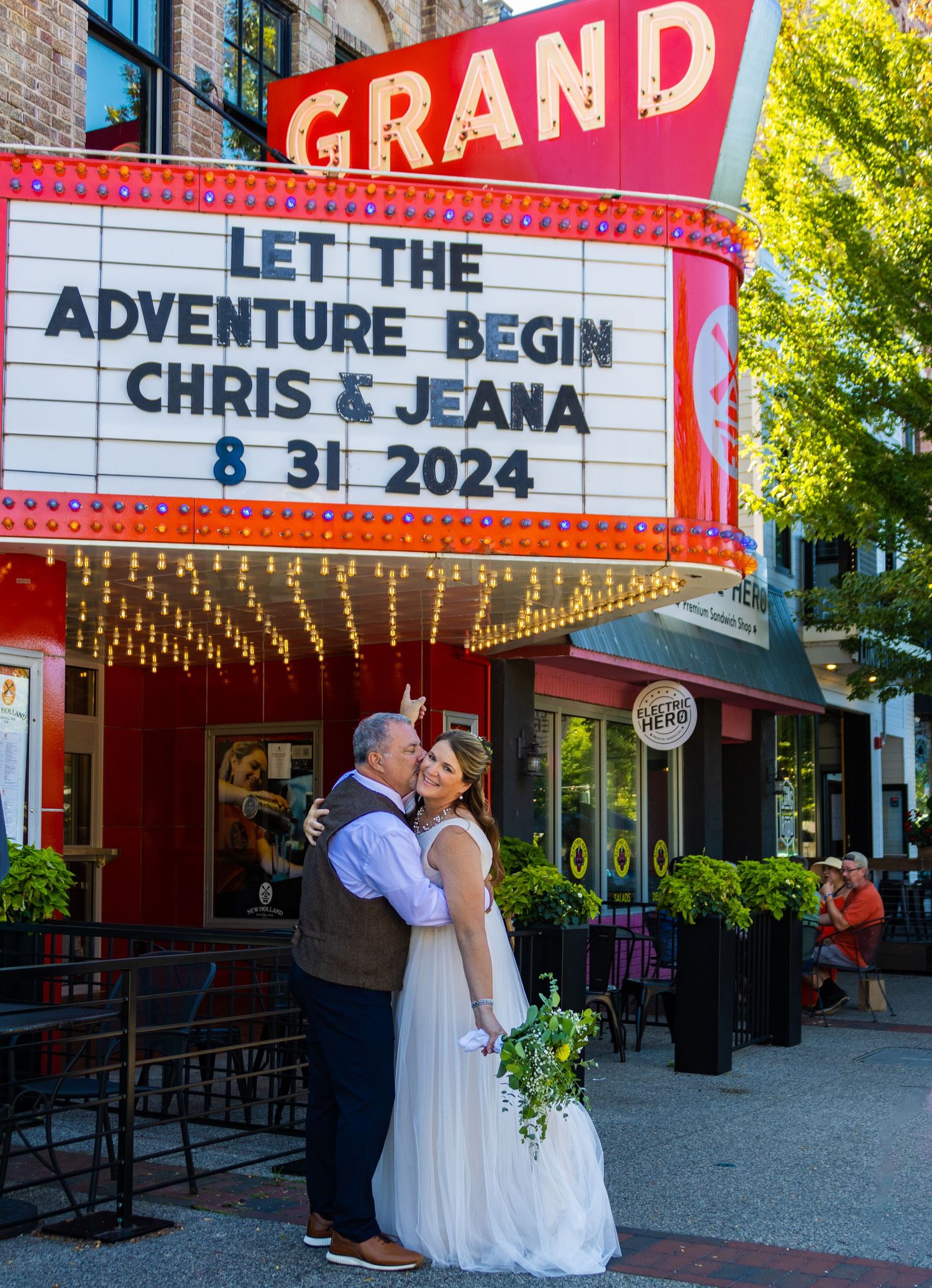 A romantic couple poses outside a vintage movie theater with a bright red marquee and Grand sign on a sunny day.