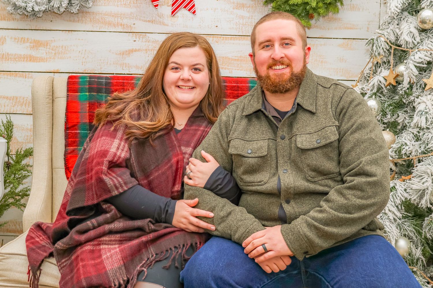 Couple sitting on bench against rustic holiday backdrop with Christmas decorations and wreaths.