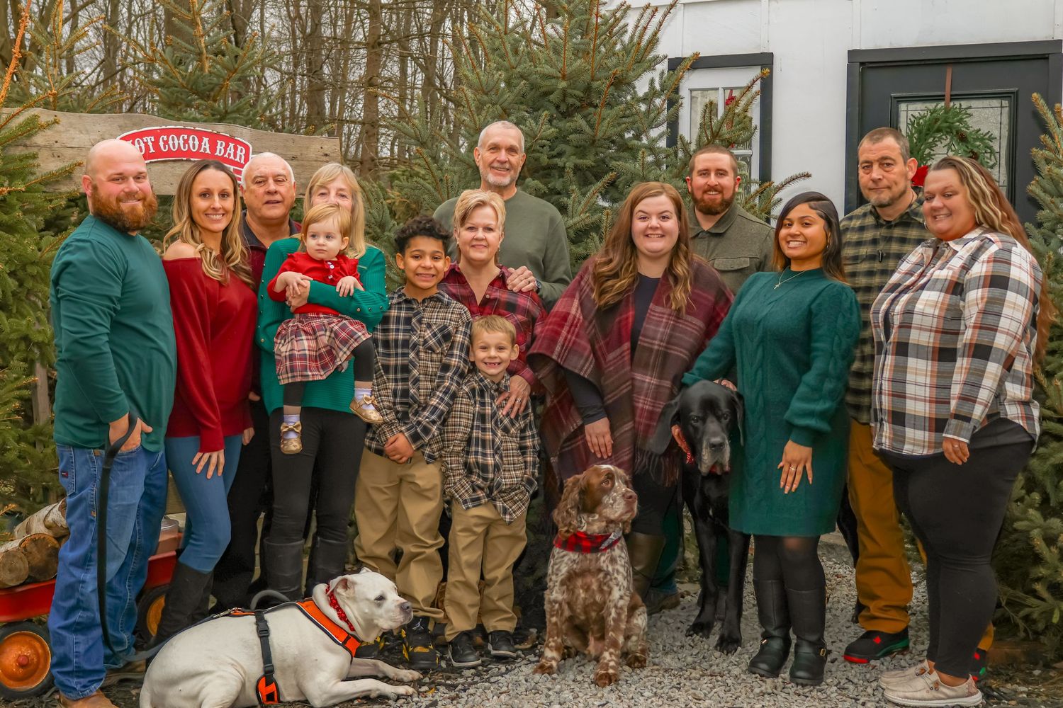 Large family group poses outdoors at Christmas tree farm with two dogs and winter decorations.