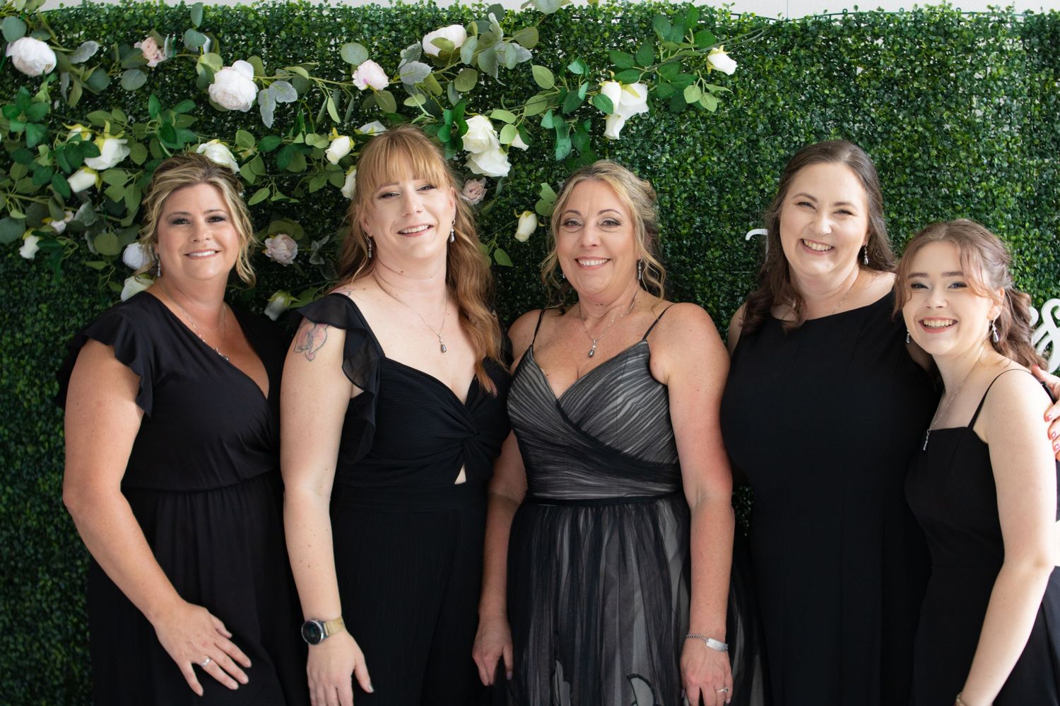 Five attendees in formal attire pose together against a green floral wall backdrop at an elegant event.