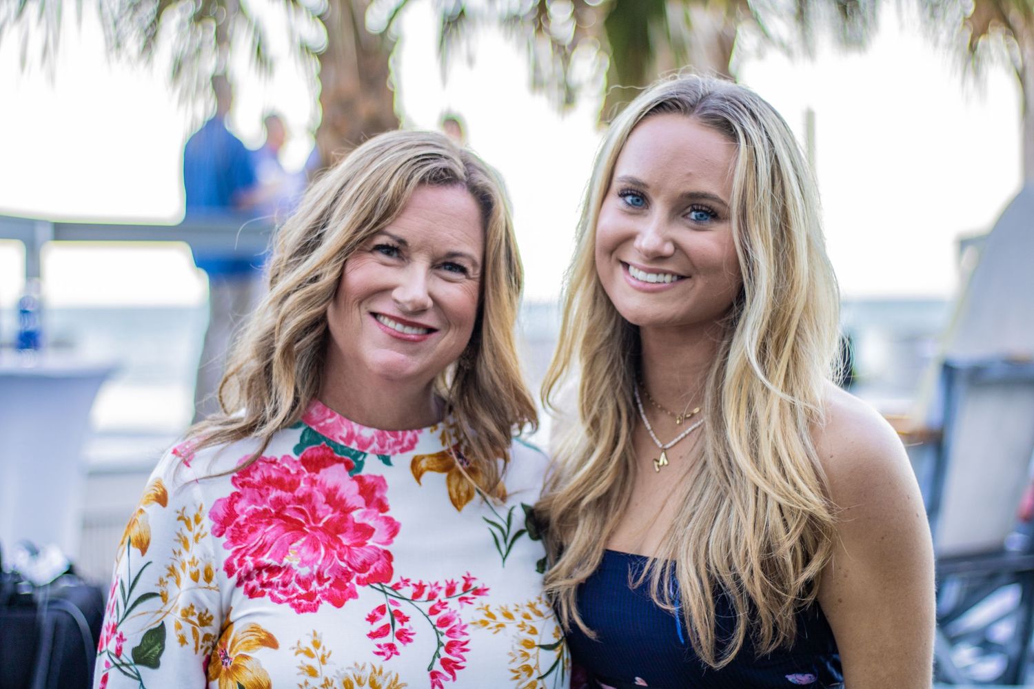 Two guests pose together at an outdoor event with palm trees in the background.