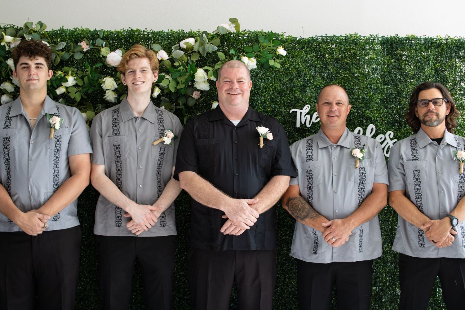 Wedding party members lined up against green hedge wall wearing coordinated attire.