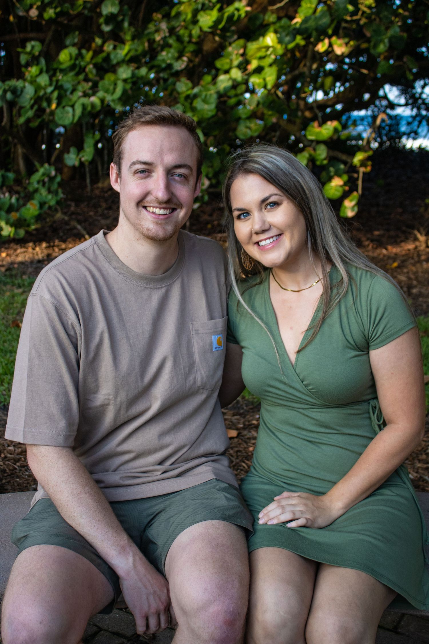 Couple in casual attire smiling while sitting on bench with fall foliage behind them.
