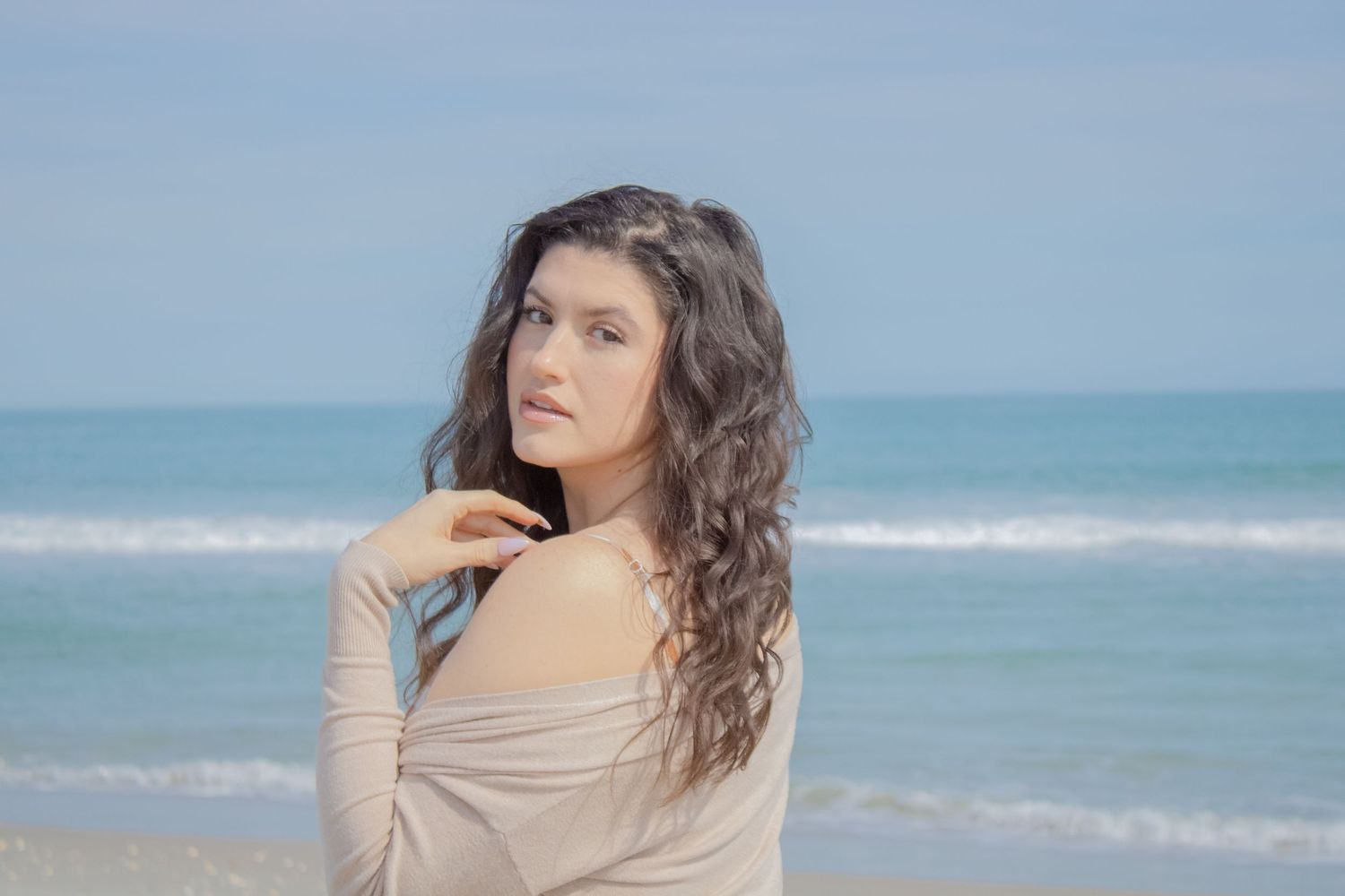 Woman with long wavy brown hair looking thoughtful on a beach with ocean in background.
