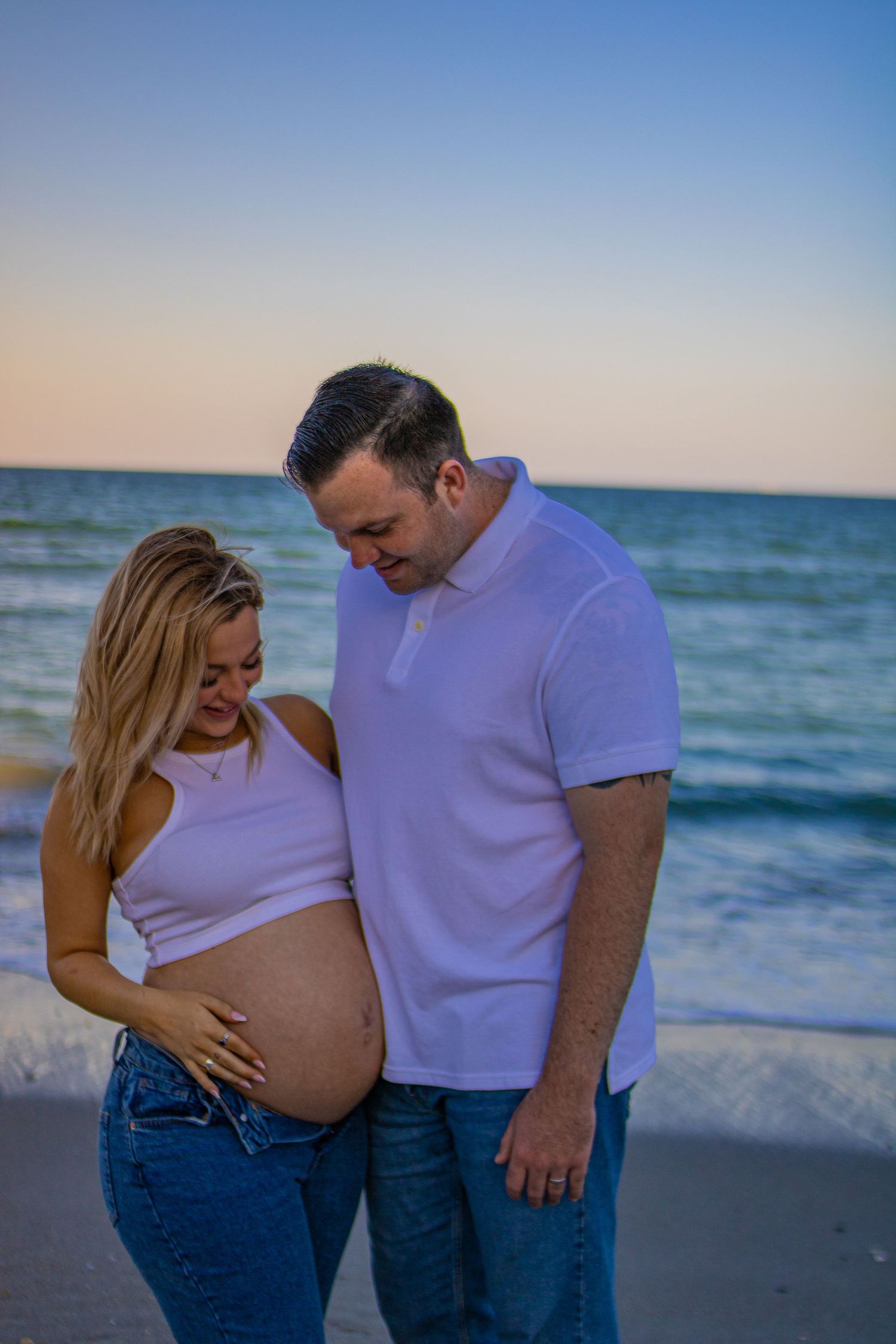 Expecting couple looks lovingly at baby bump during sunset beach photoshoot.