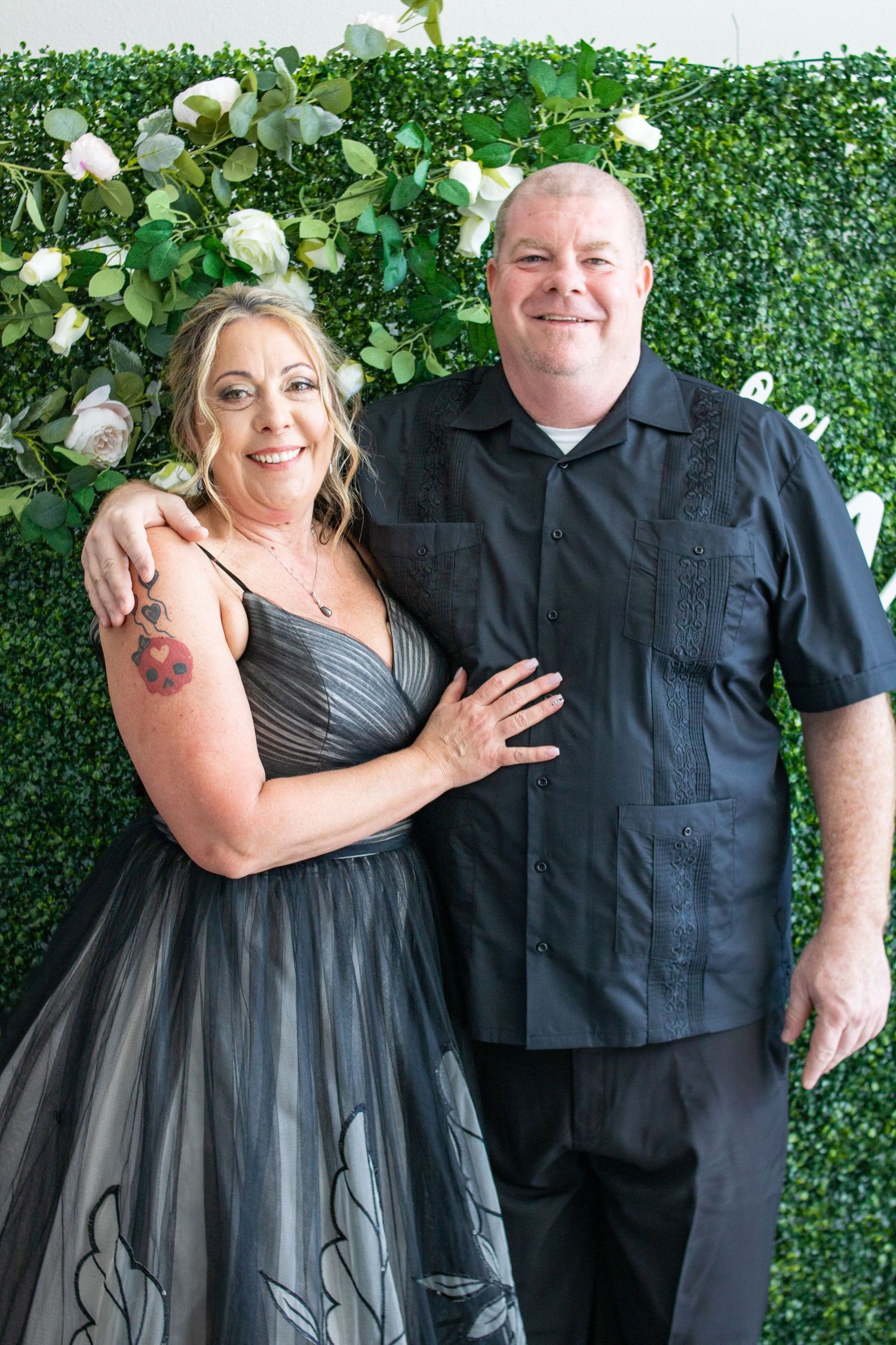 Joyful couple poses against green wall with white roses for formal portrait.