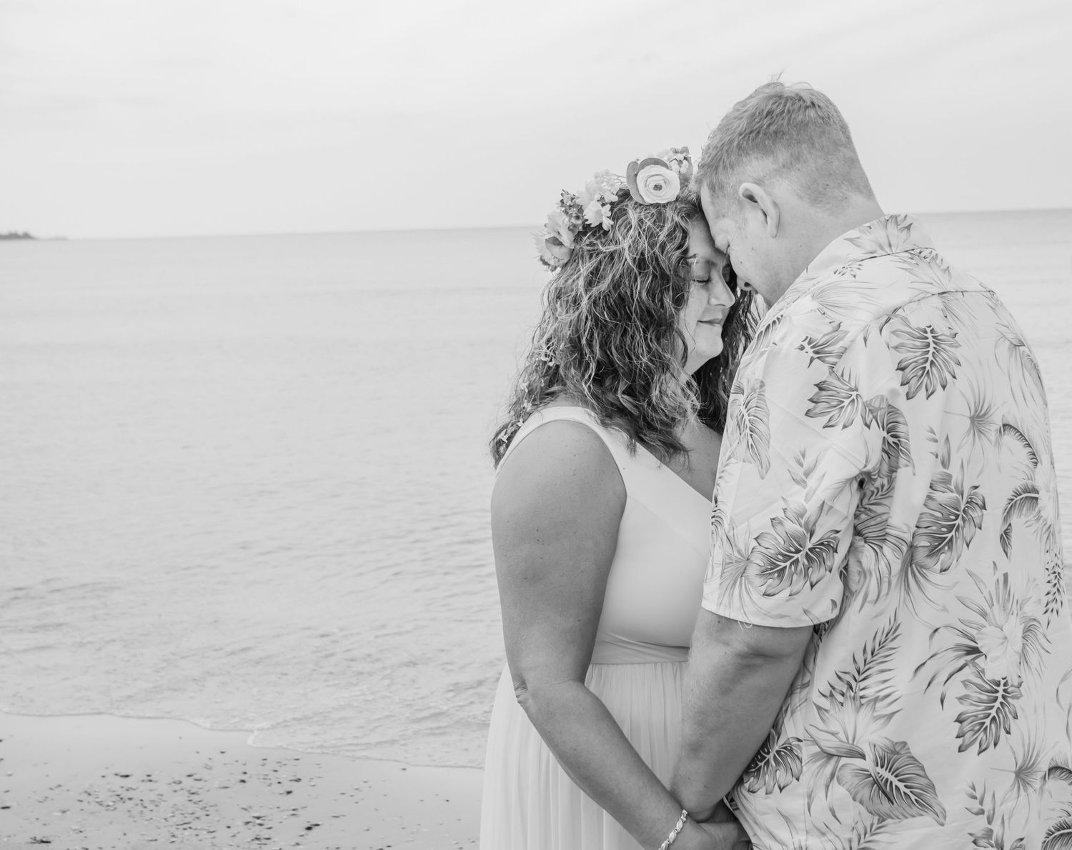 A romantic couple embraces on a sandy beach at sunset while wearing casual summer attire.