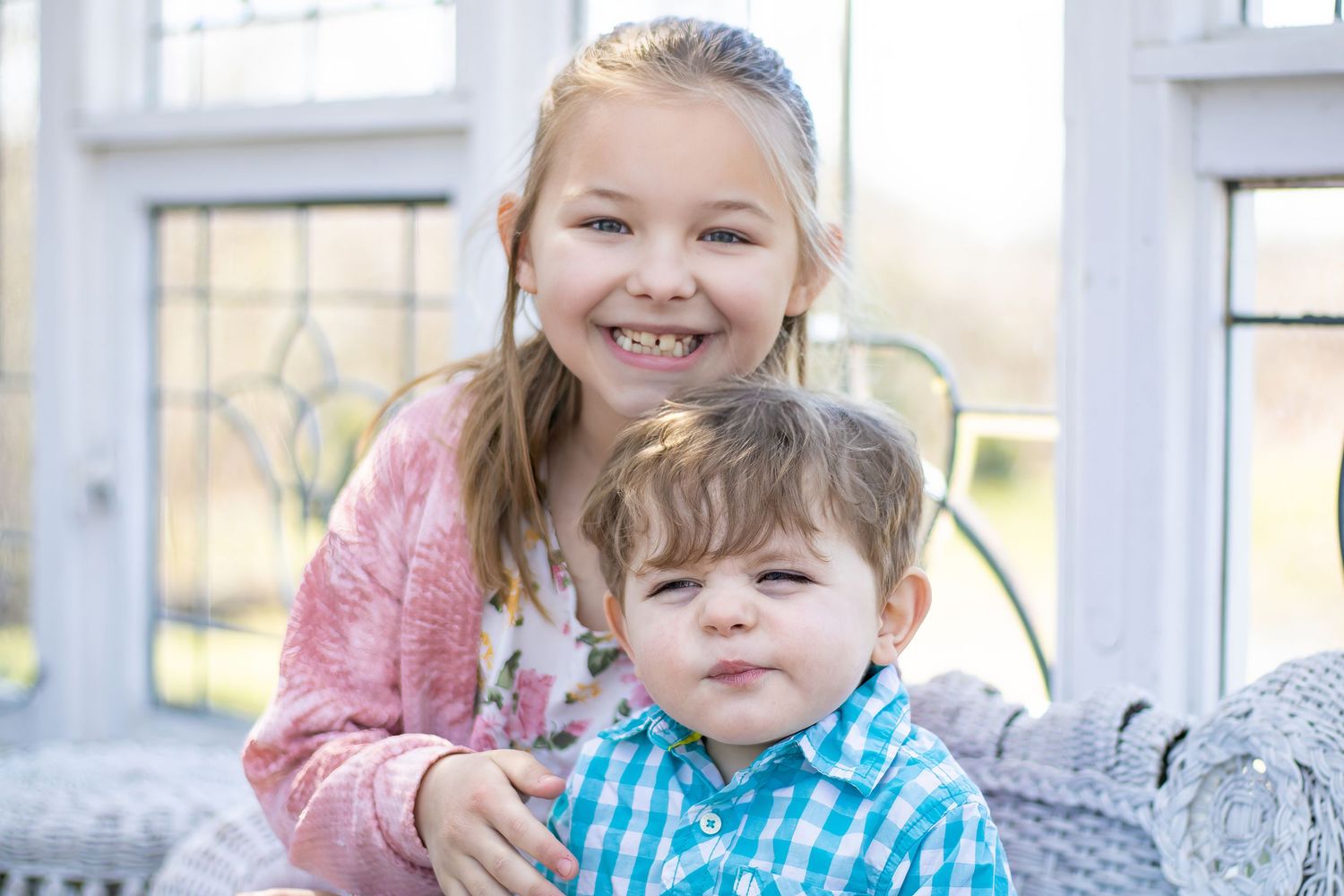 Two siblings sitting together on a white bench with sunny windows in the background.