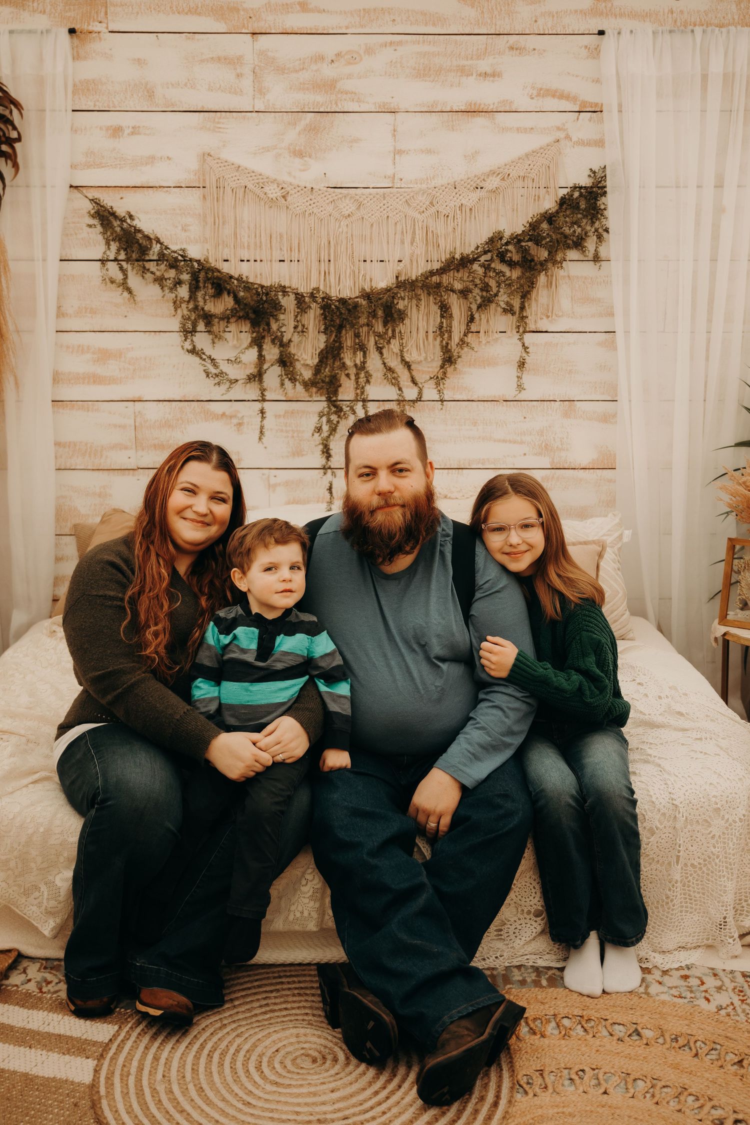 Family of four poses together on white bed with macrame wall hanging decoration.