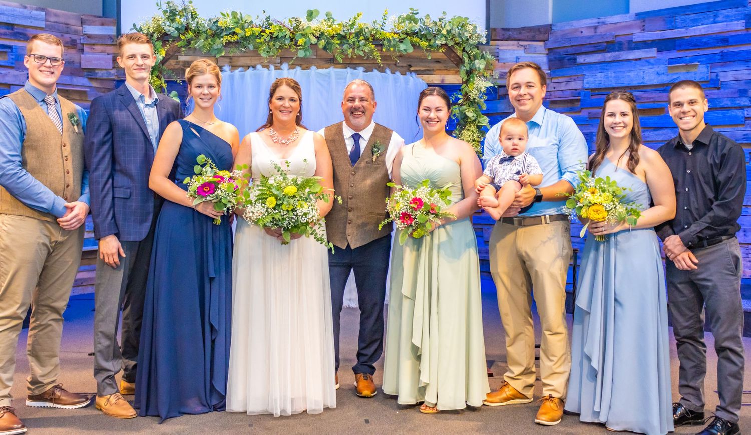 Large wedding party group photo with mix of formal attire under decorated arch with blue backdrop.