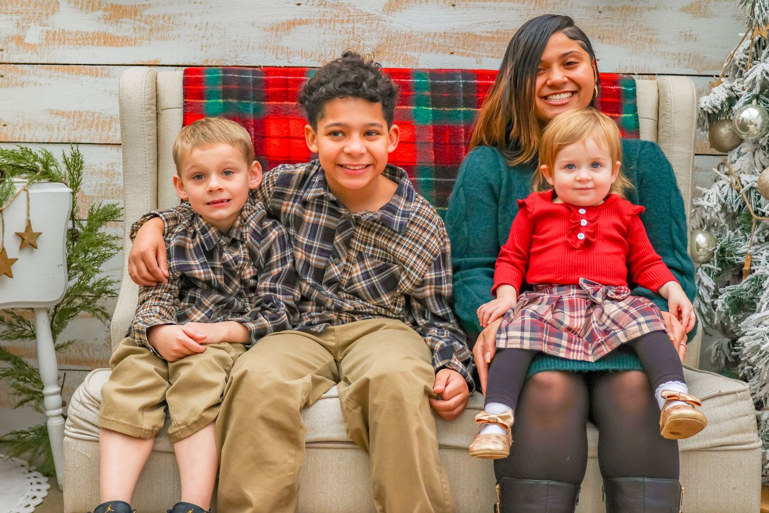 A festive holiday portrait on a red and green plaid bench with whitewashed wood background and evergreen decorations.