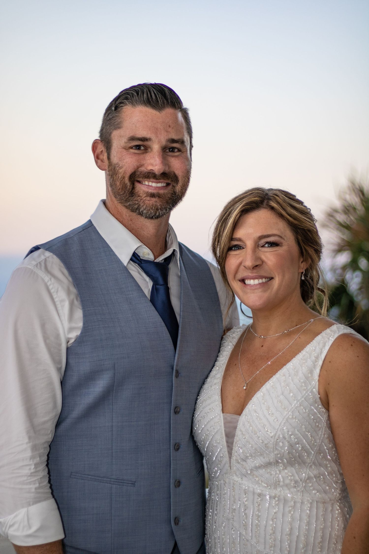 A bride and groom pose together outdoors at dusk in formal wedding attire.
