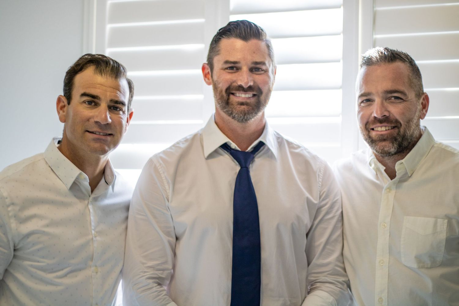 Three men in formal attire smiling indoors at a wedding.