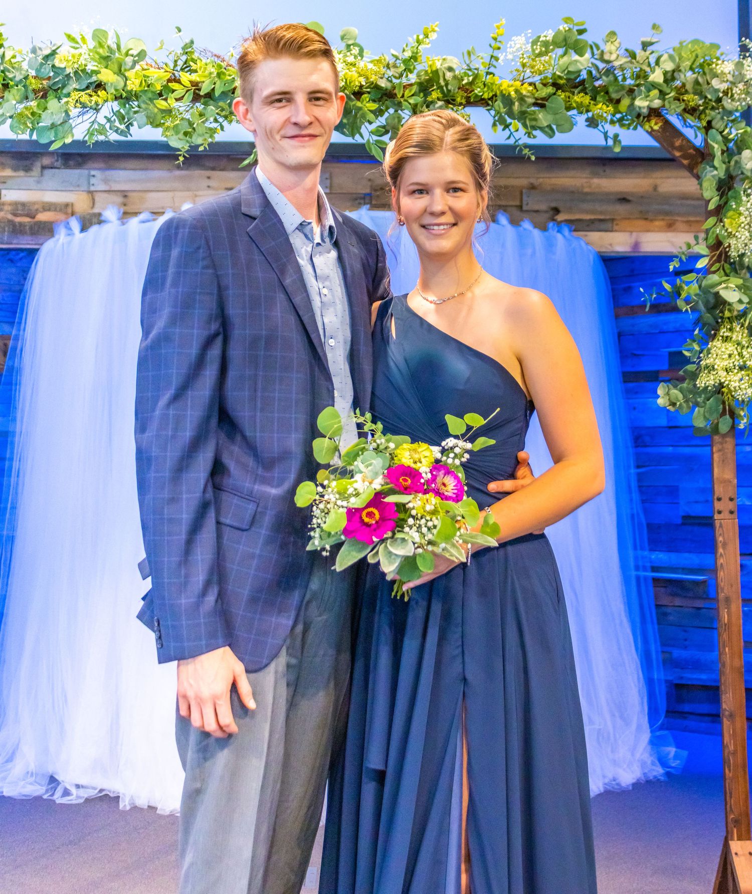 A couple poses beneath a floral arch decorated with blue drapery at a formal event.