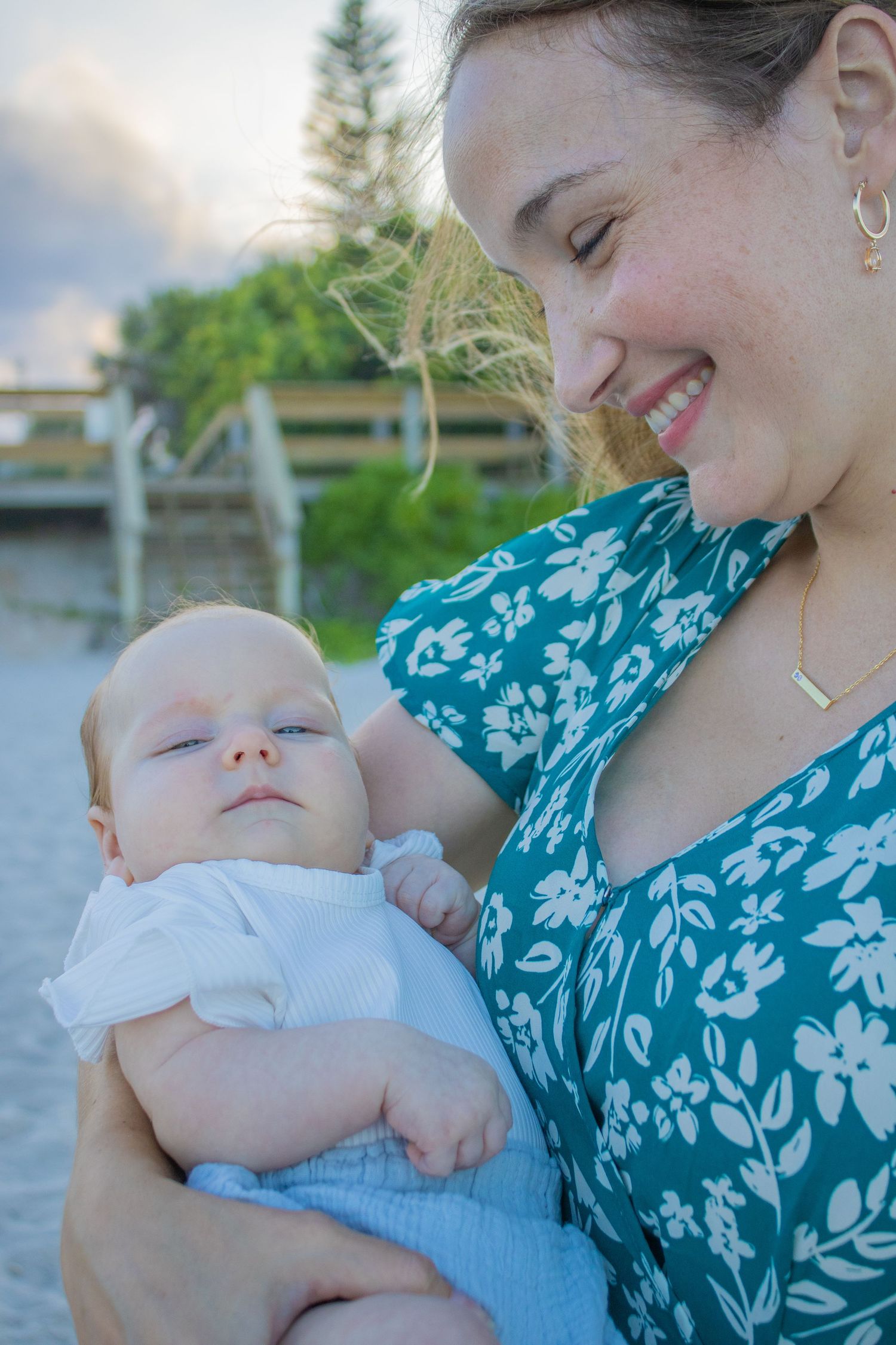 A tender moment shows a baby resting peacefully against someone wearing a floral teal dress outdoors.