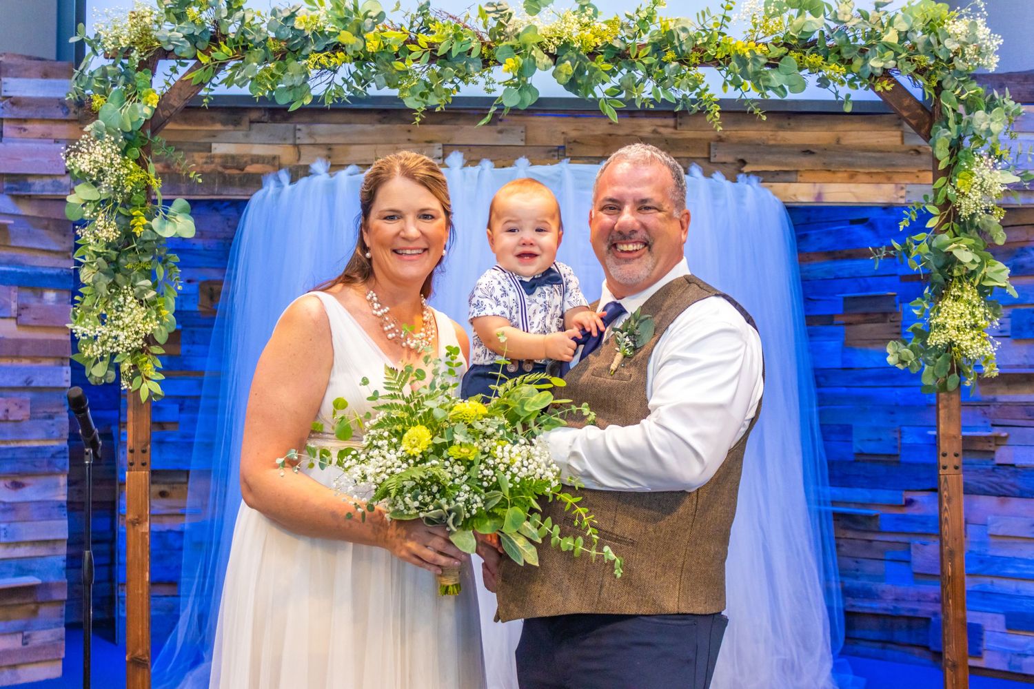 Wedding portrait under floral arch with blue backdrop featuring couple and young child.