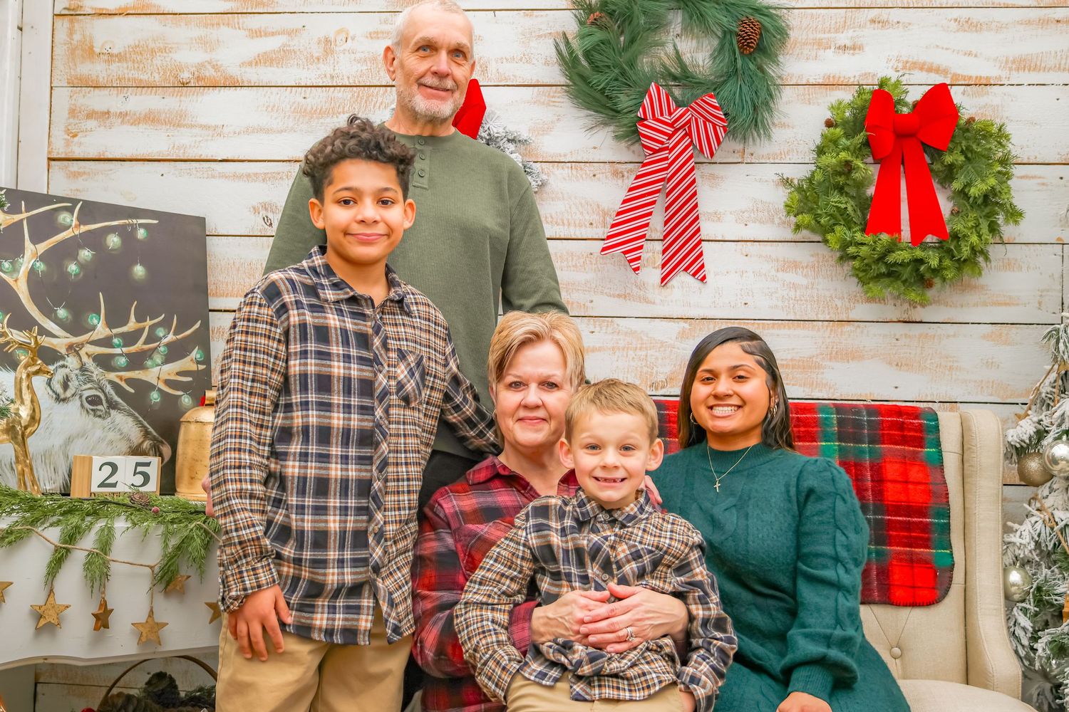 A family poses together in front of a rustic white wall decorated with Christmas wreaths and bows.