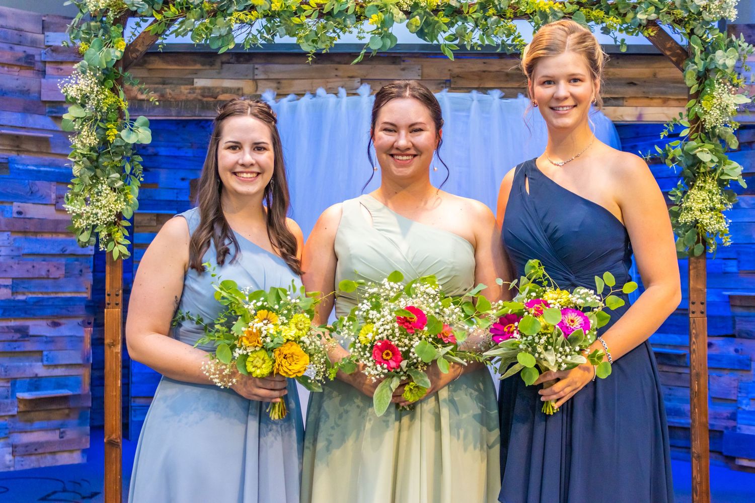 Three attendants pose in formal dresses holding colorful bouquets against a blue backdrop with floral arch.
