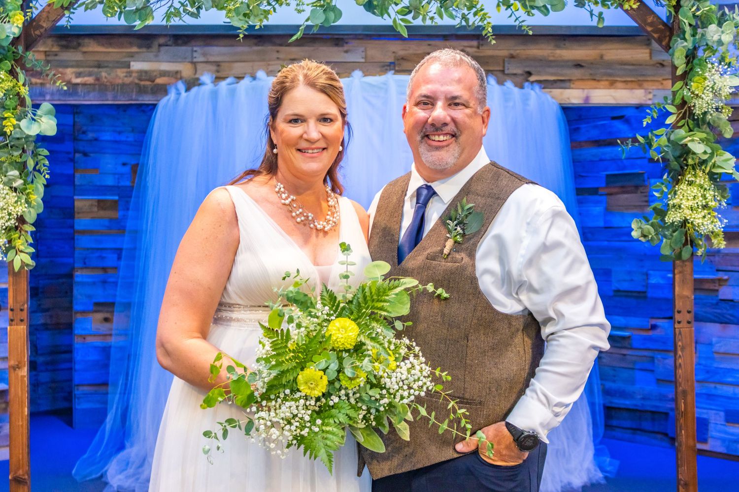 Couple poses together under floral arch with bright blue backdrop at wedding celebration.