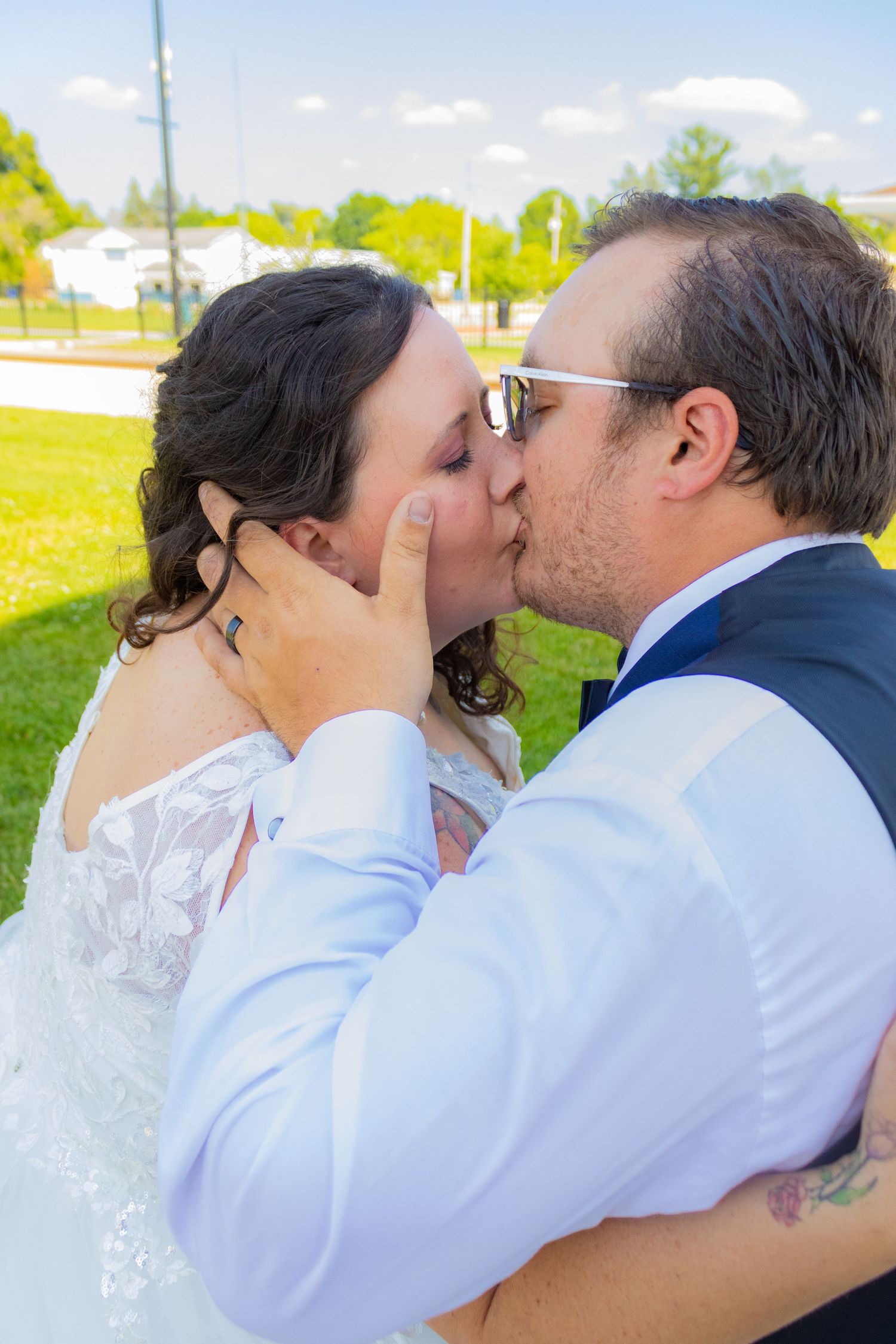A couple in wedding attire share a romantic kiss outdoors on a sunny day with green grass and blue sky in the background.