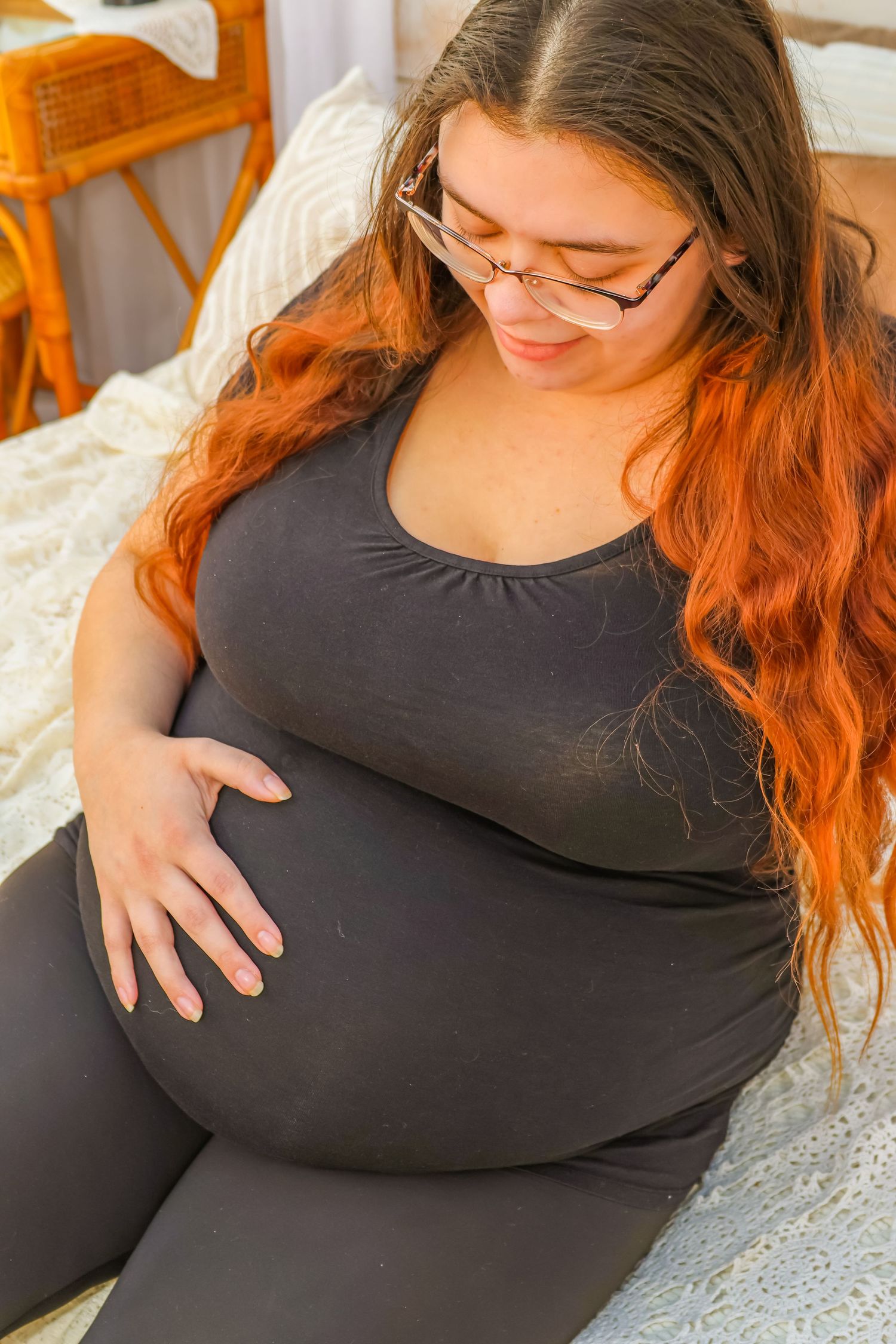 Expectant mother in black attire rests hand on belly while sitting on white bedding.