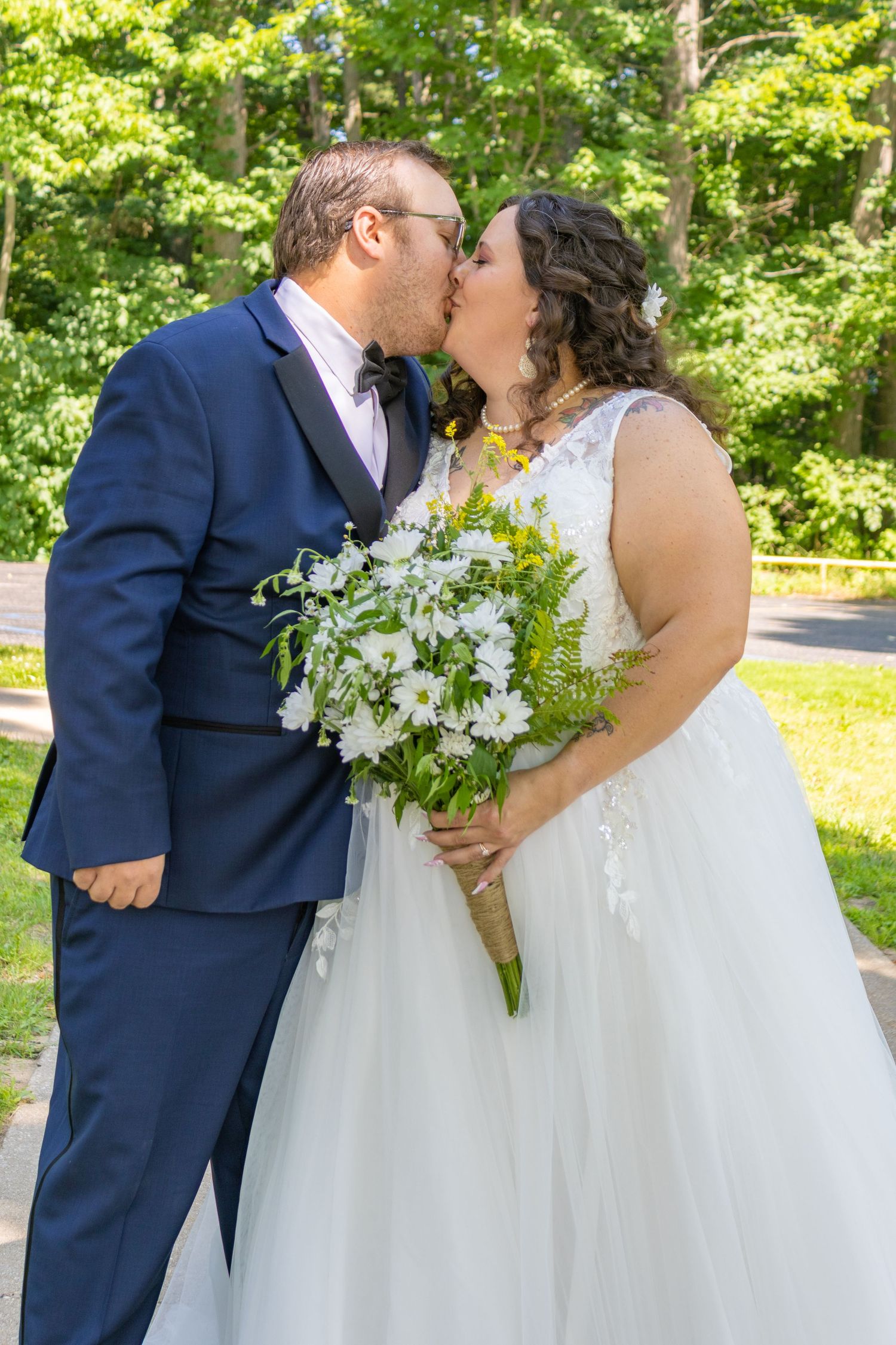 Wedding couple sharing kiss outdoors while holding white floral bouquet.
