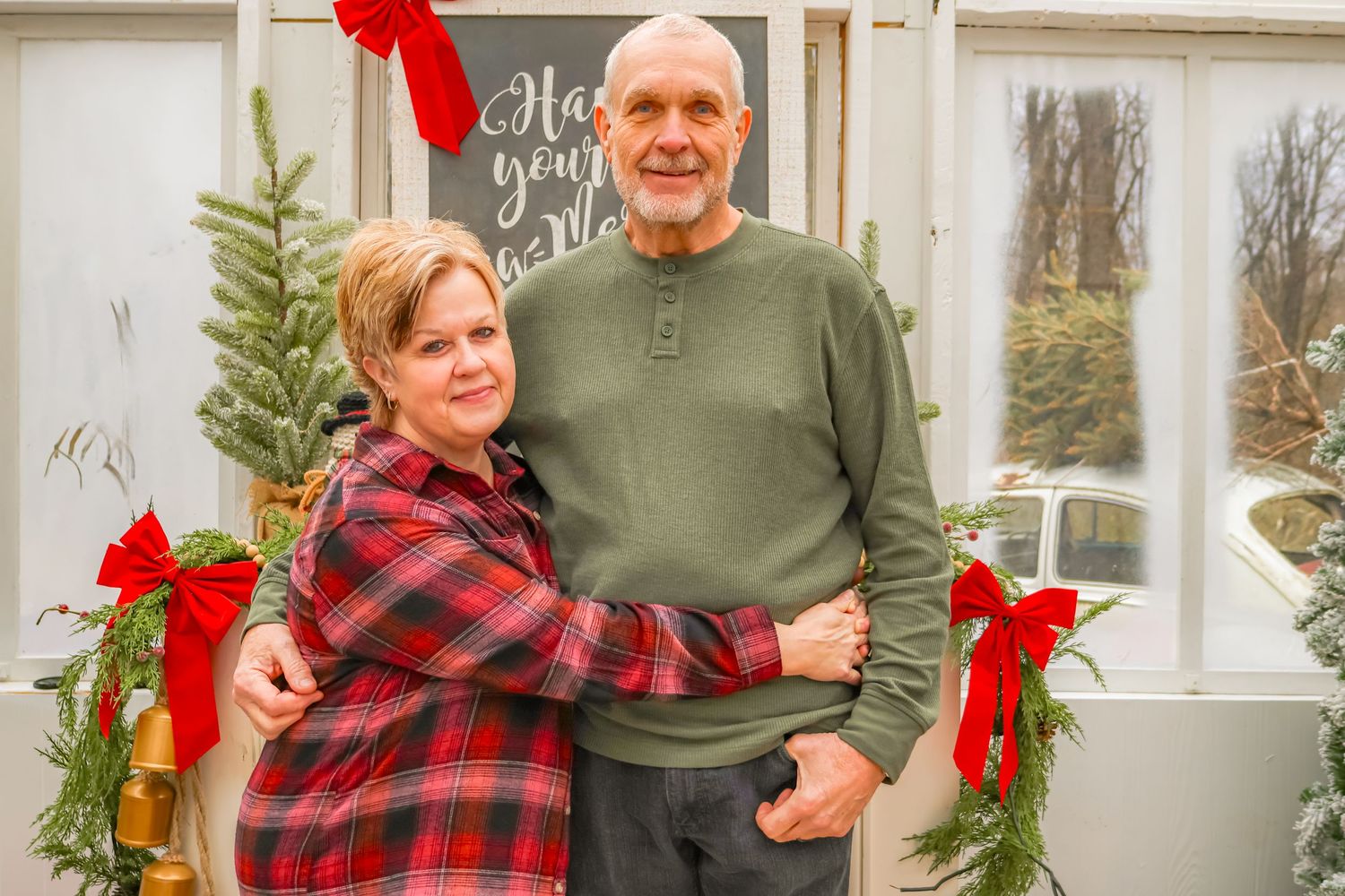 Couple in festive attire standing in front of holiday-decorated window with wreaths and greenery.