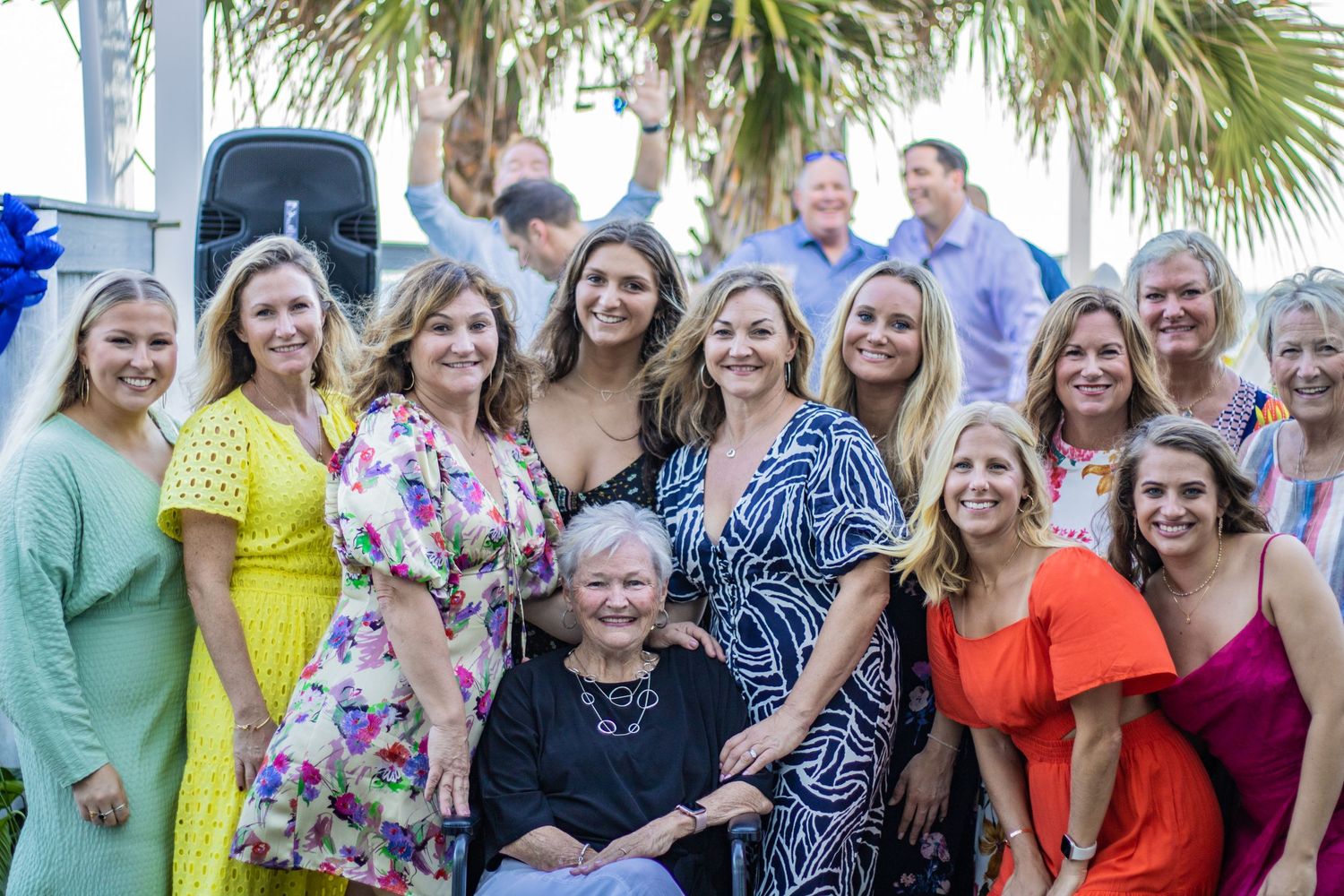 A group photo of guests in colorful dresses gathered outdoors with palm trees in the background at a celebration.