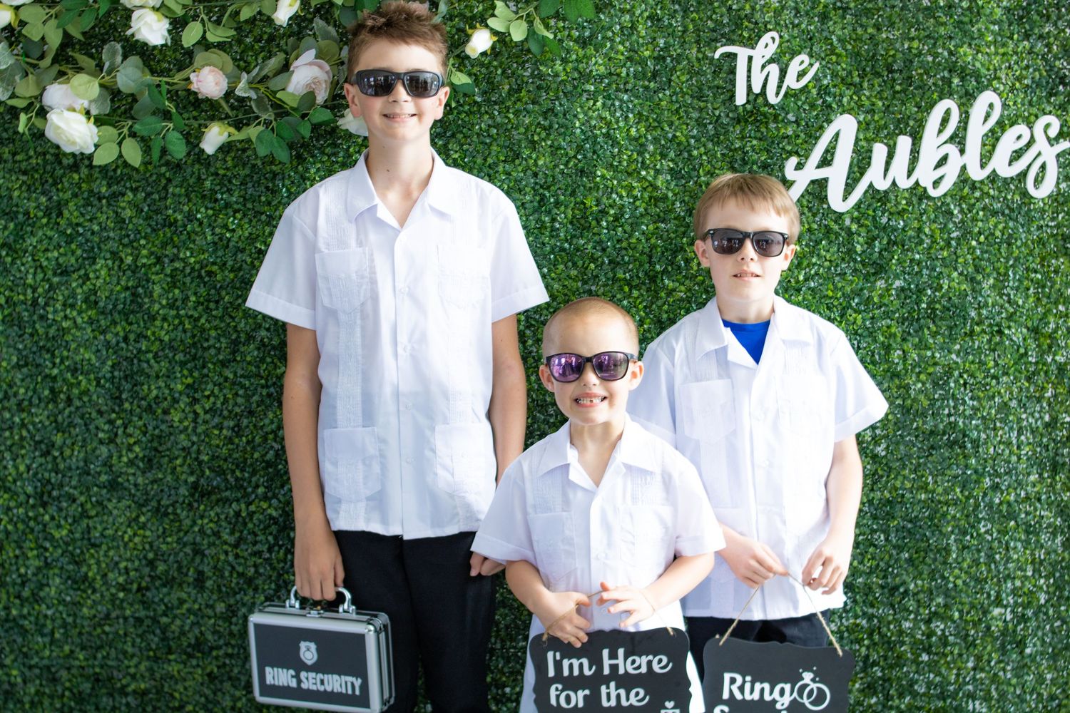 Three young attendants in white shirts and sunglasses pose against green hedge wall.