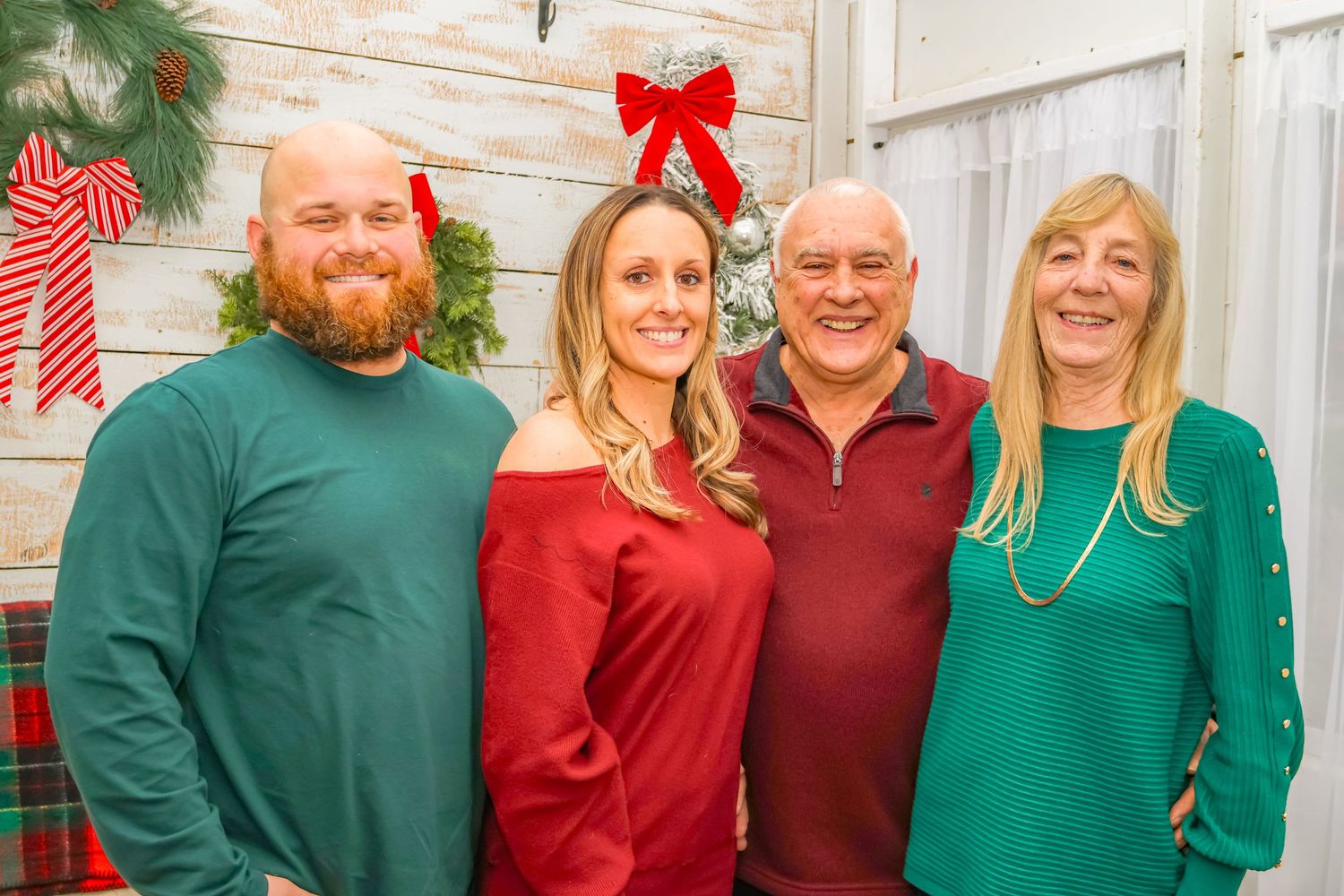 Group portrait in festive attire against white wooden wall with Christmas greenery and red bow.