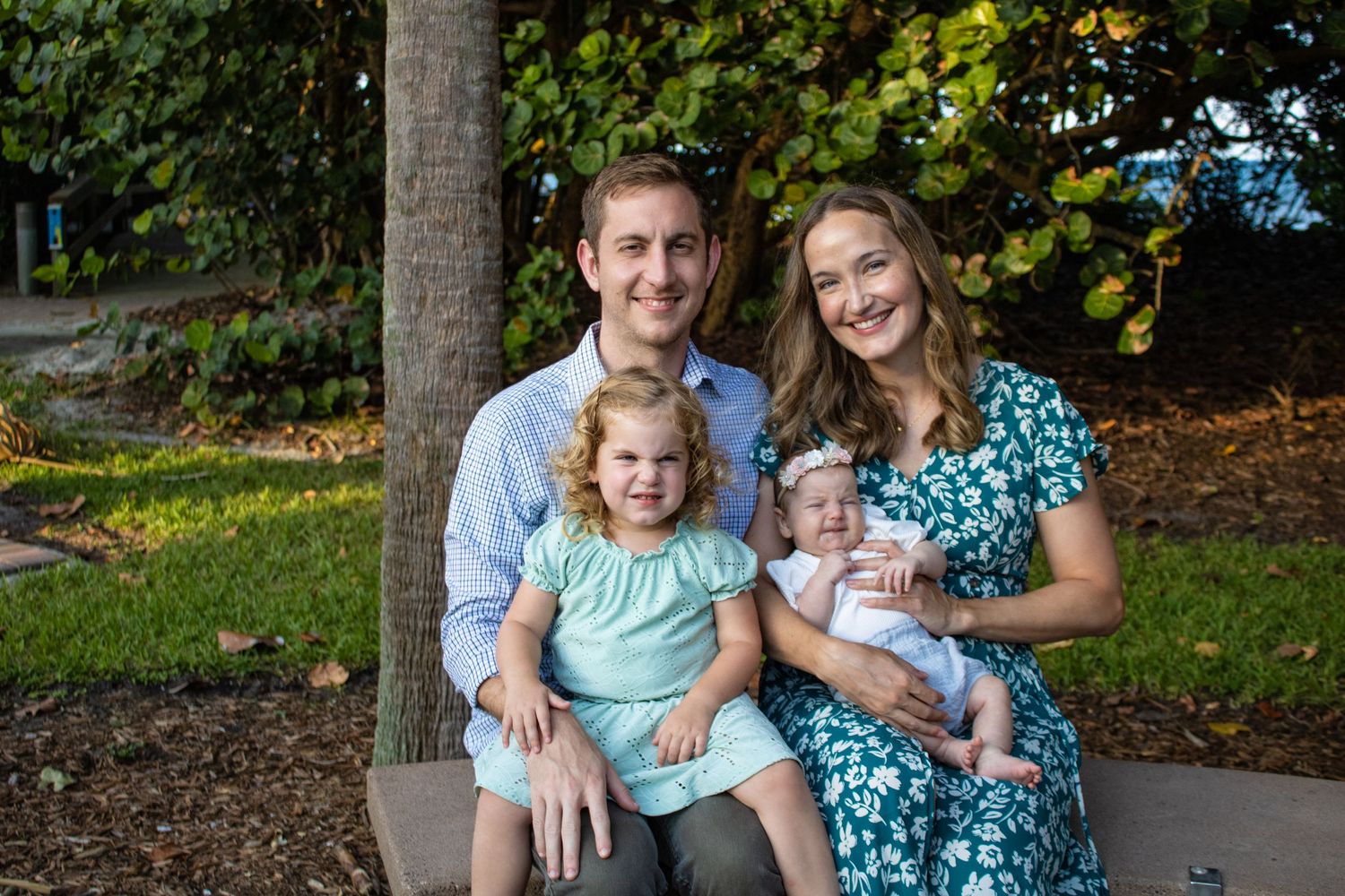 Family of four sits together outdoors in casual clothing near a tree.