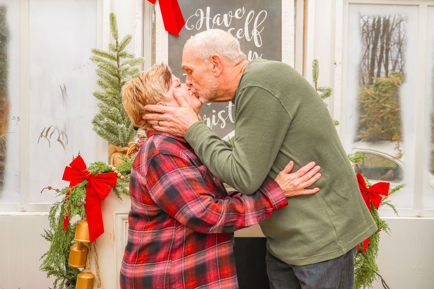 A couple embraces in front of festive holiday decorations with wreaths and red bows.