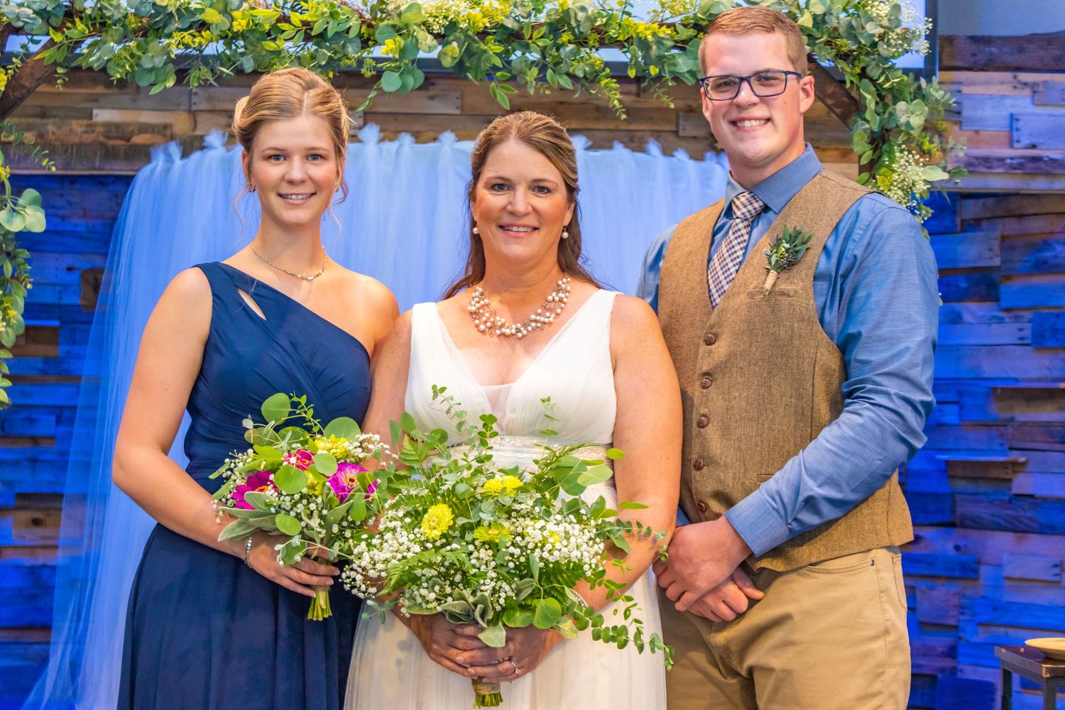 Three people pose under a floral arch with greenery and blue draping at a wedding ceremony.