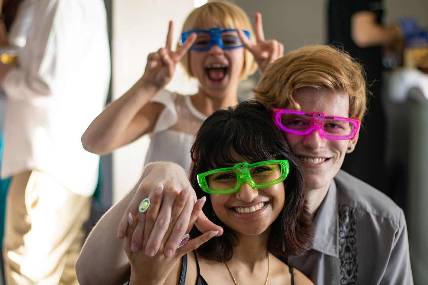 Party guests wearing colorful novelty sunglasses celebrate while showing off an engagement ring.