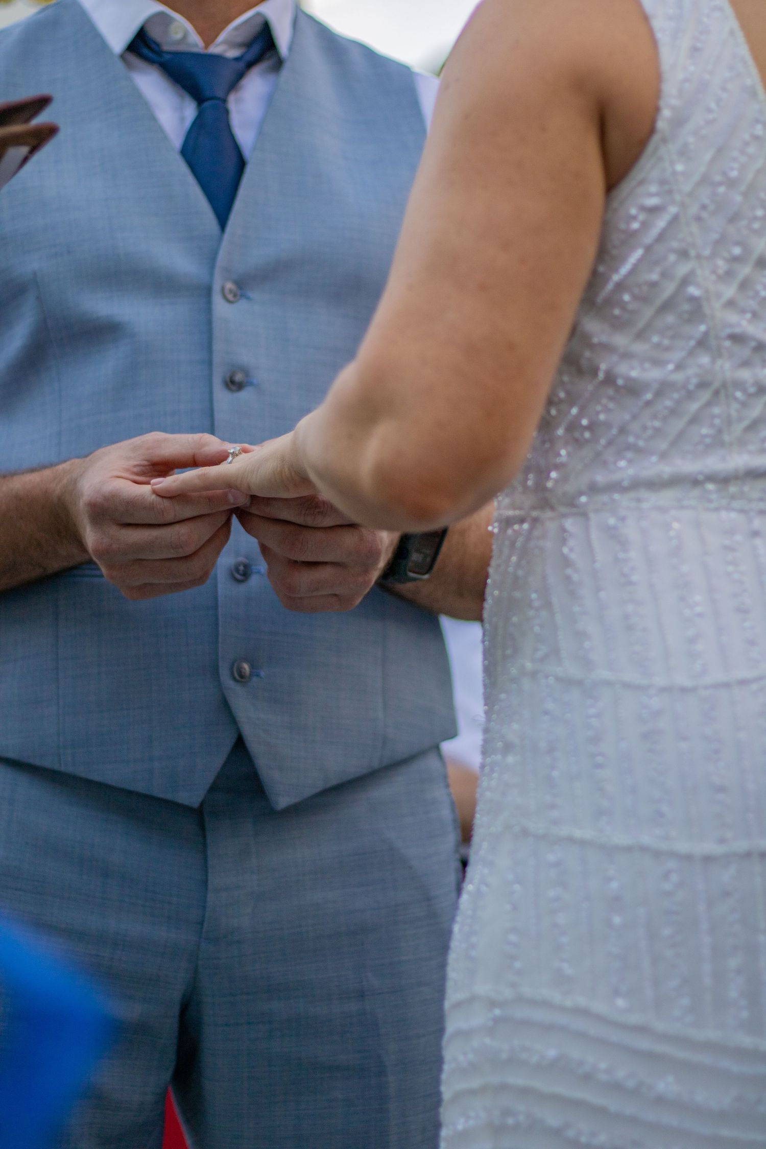 Close-up of couple exchanging rings during wedding ceremony with bride in white dress and groom in blue vest.