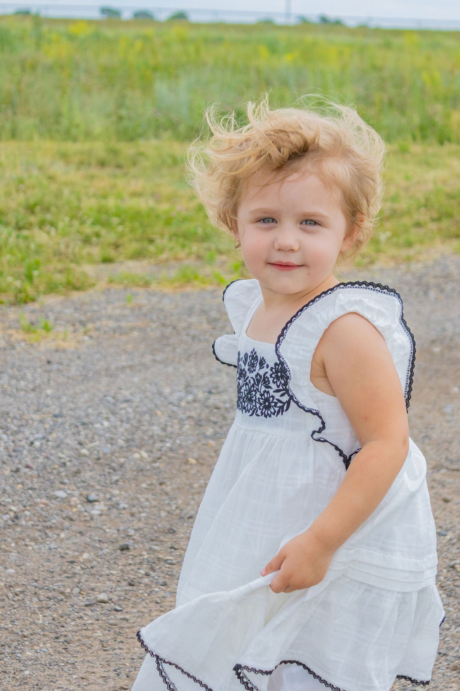 Young child in white dress with navy trim smiles while standing on dirt path.