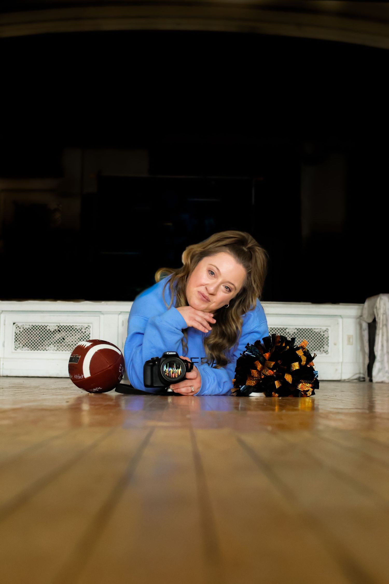 Person in blue sweater lies on wooden floor with football and cheerleading pom poms.