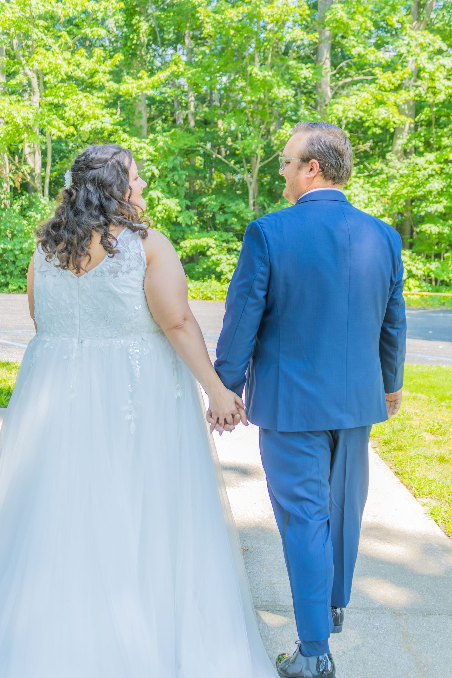 Newlyweds walking hand in hand on a path, bride in white dress and groom in blue suit.