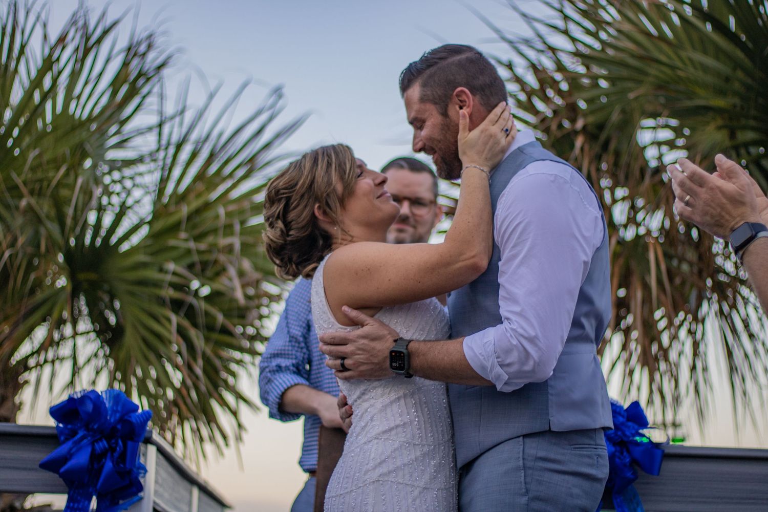 Newlyweds share their first kiss at outdoor wedding ceremony with palm trees in background.