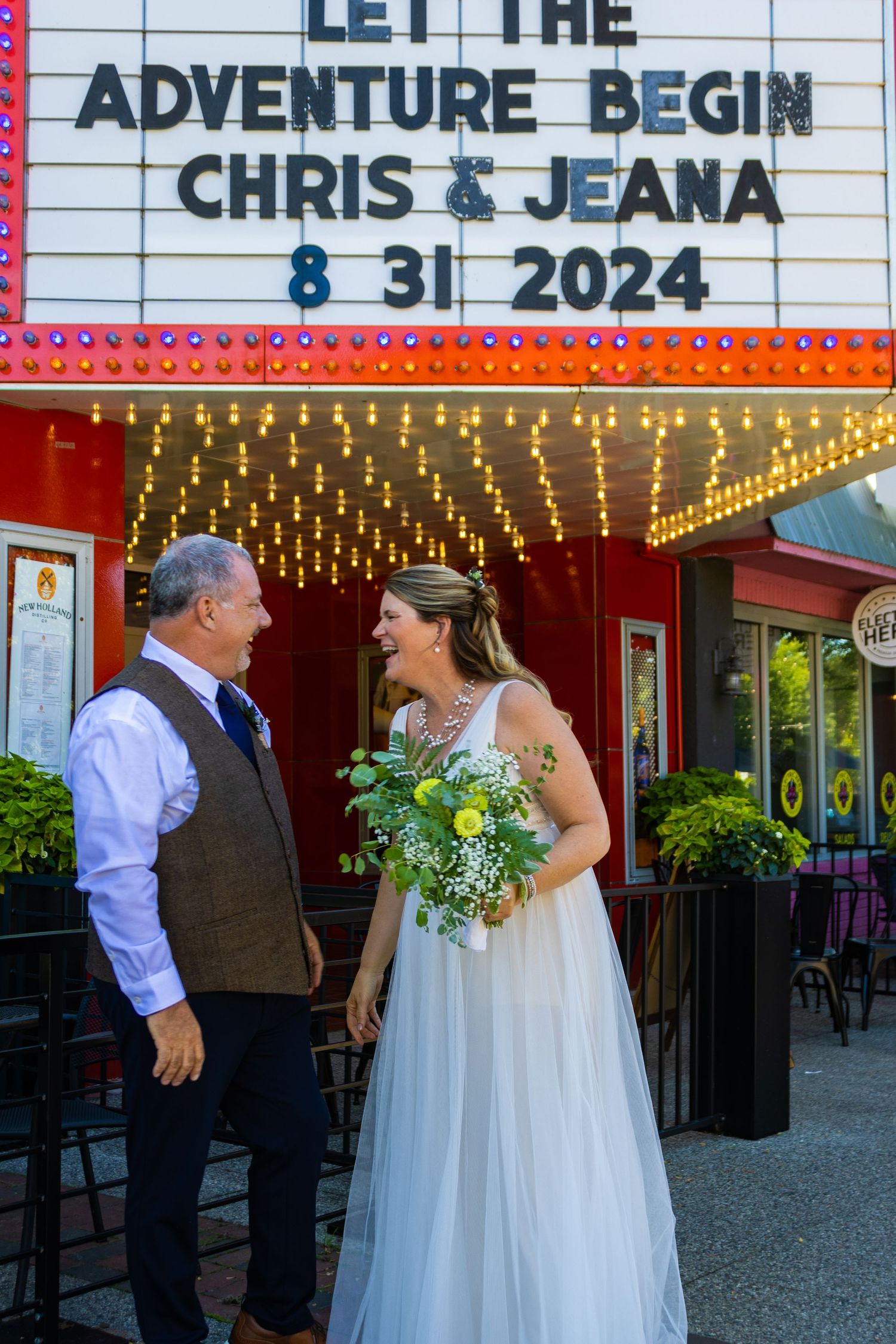 Newlyweds share intimate moment under illuminated theater marquee displaying wedding details.