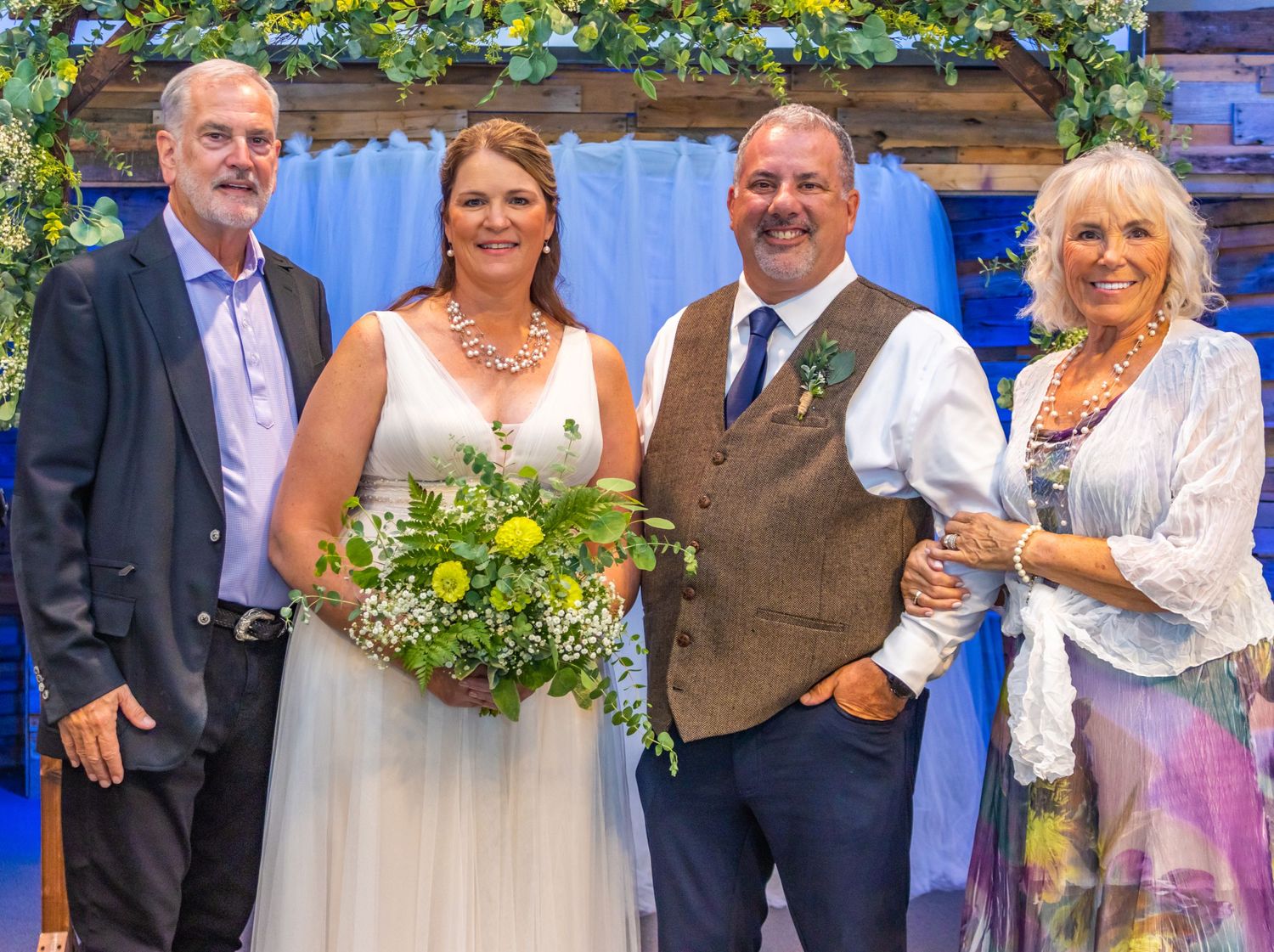 Wedding group portrait in front of blue backdrop with floral arch decoration overhead.
